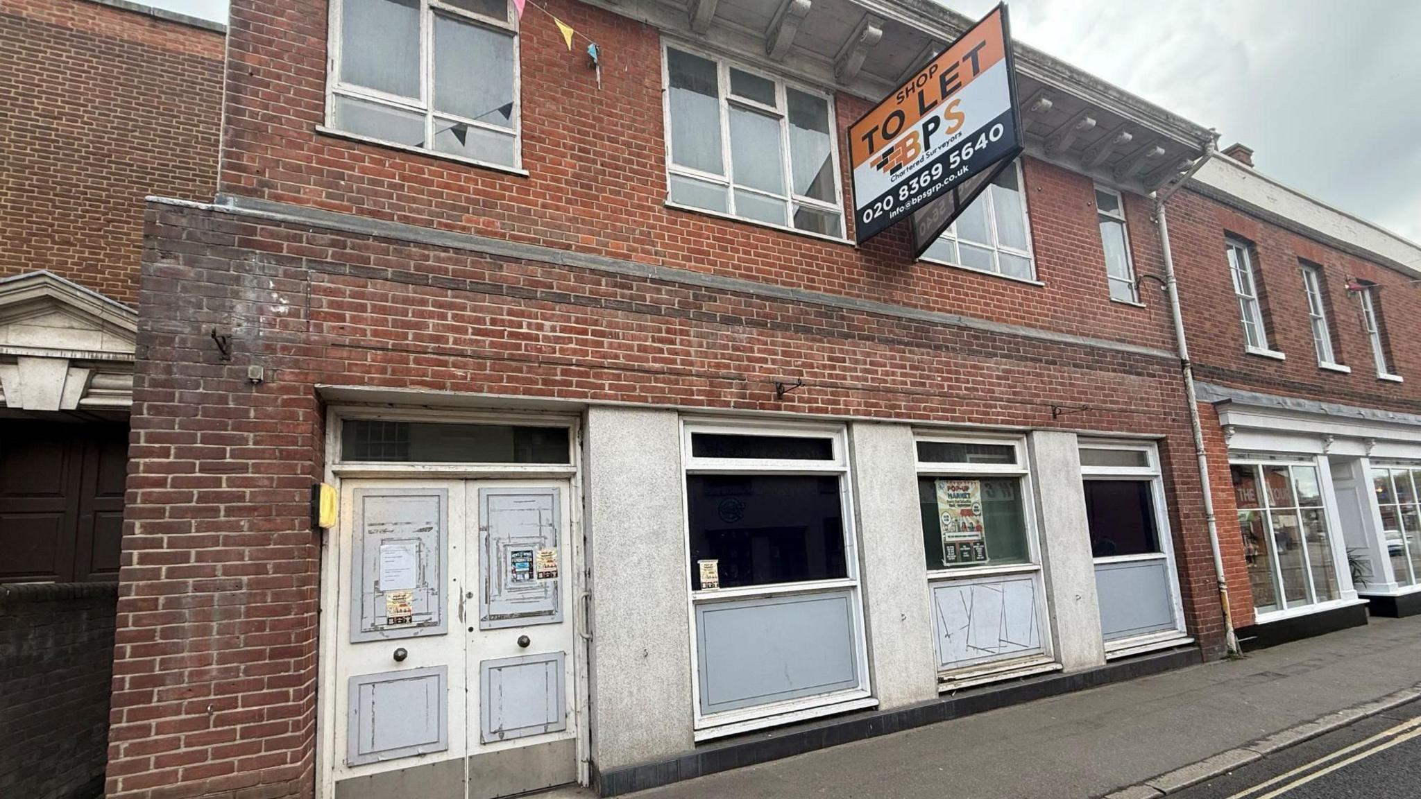 The derelict former Barclays bank, which has a brick facade with faded white columns. The door and windows look aged and are slightly cracked. There is an orange, white and black to let sign on the building.