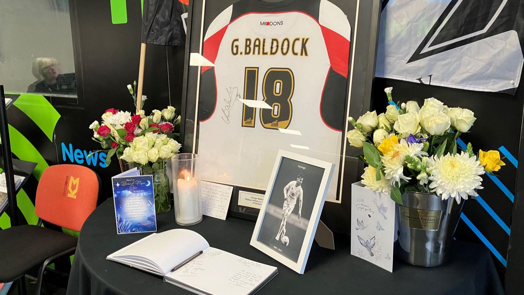 A framed football shirt with the name G. Baldock and the number 18, is on a table alongside a condolences book, candle, cards and vases of white, red and yellow roses and white chrysanthemums. A small, black and white framed photograph of the player is also on the table.