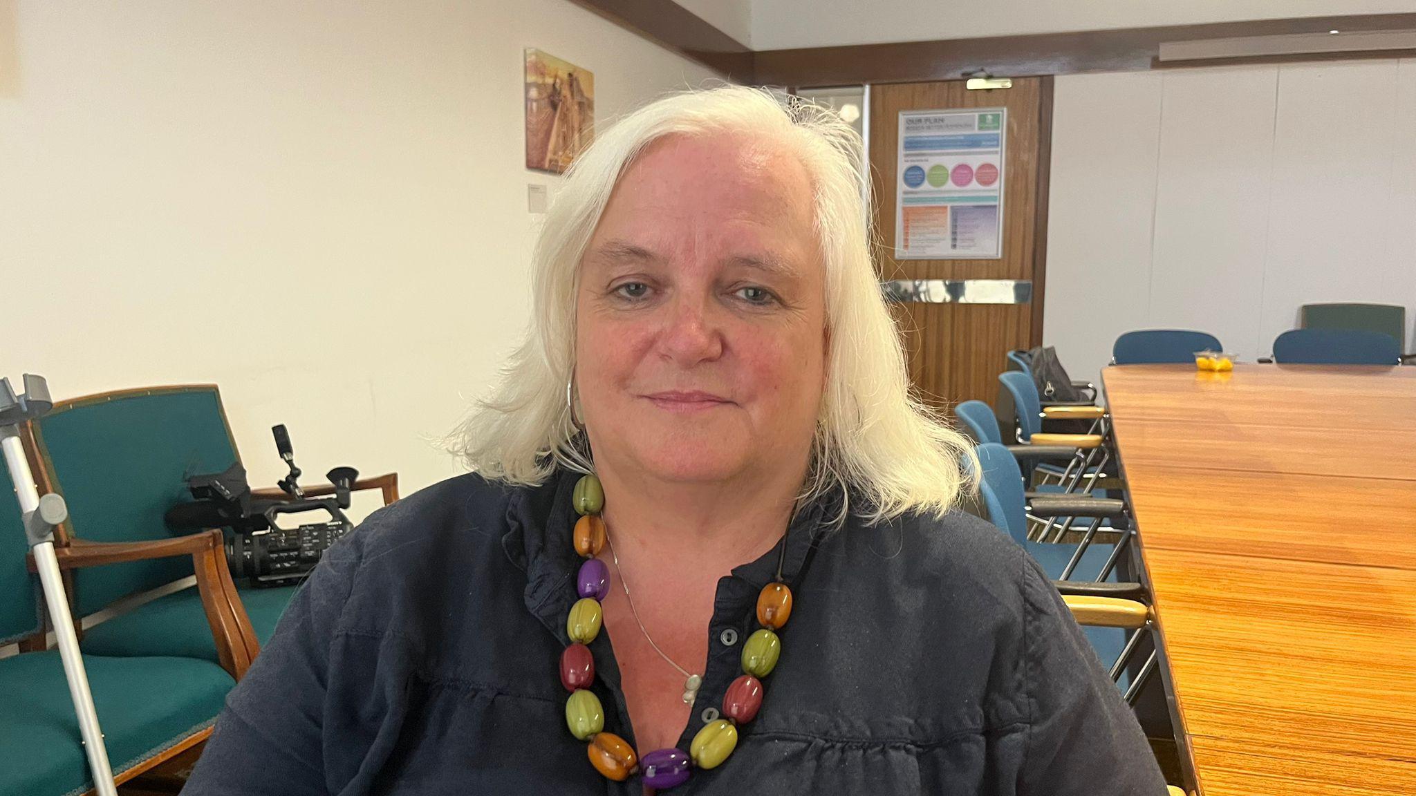 Jemima Laing wearing a dark top and a colorful beaded necklace, is seated in a room. The background includes chairs, a walking aid, a wooden table with chairs, and a poster on the wall near a door.