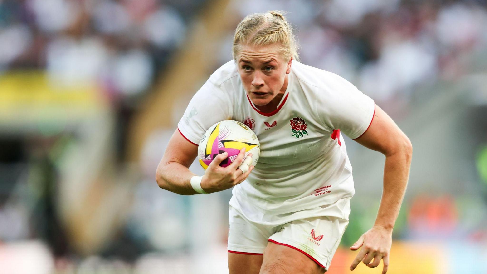 Alex Matthews runs with her body bent slightly forward and a rugby ball tucked to her chest under her right arm as she looks ahead during a World Cup match for England in 2025