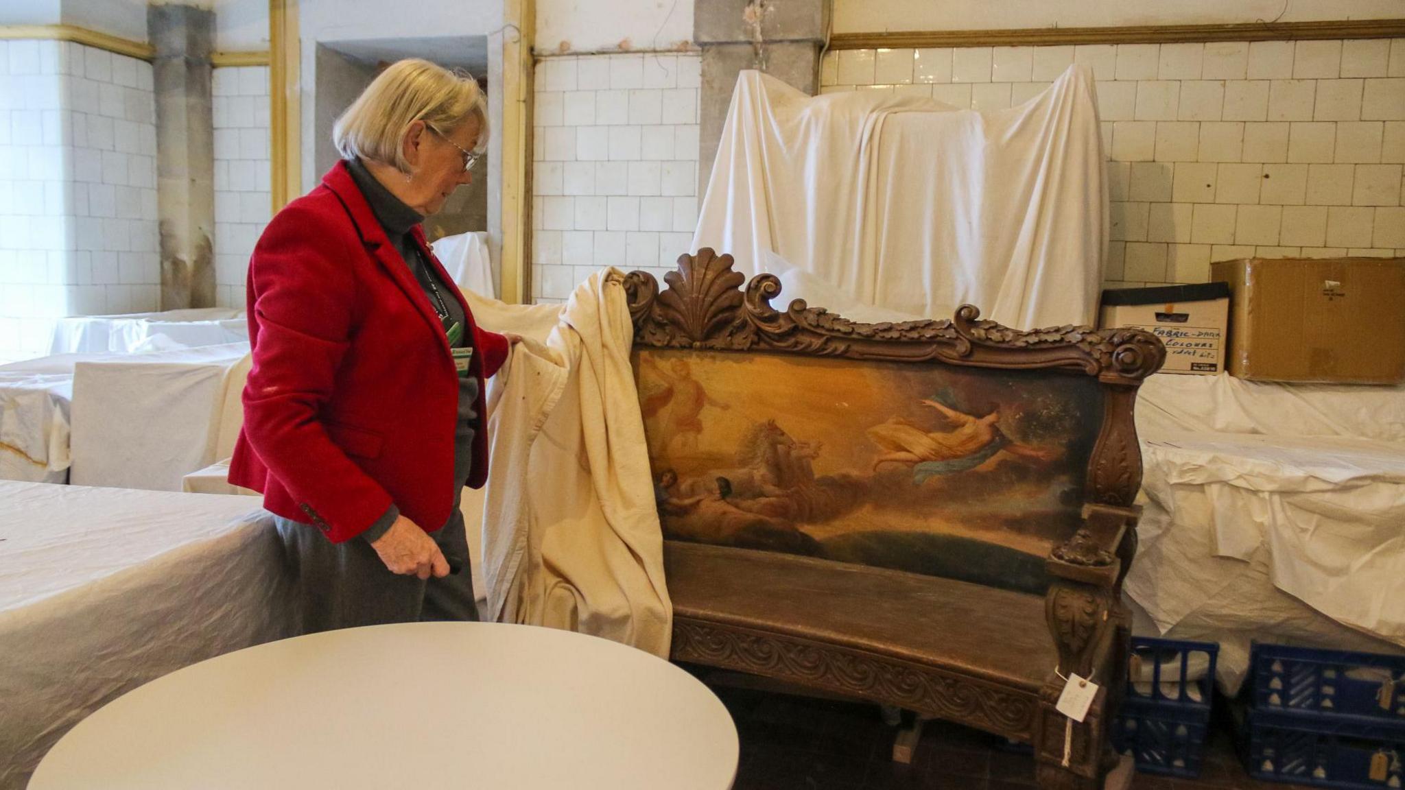 A woman wearing a red jacket uncovers a decorated bench in a room with various other covered objects 