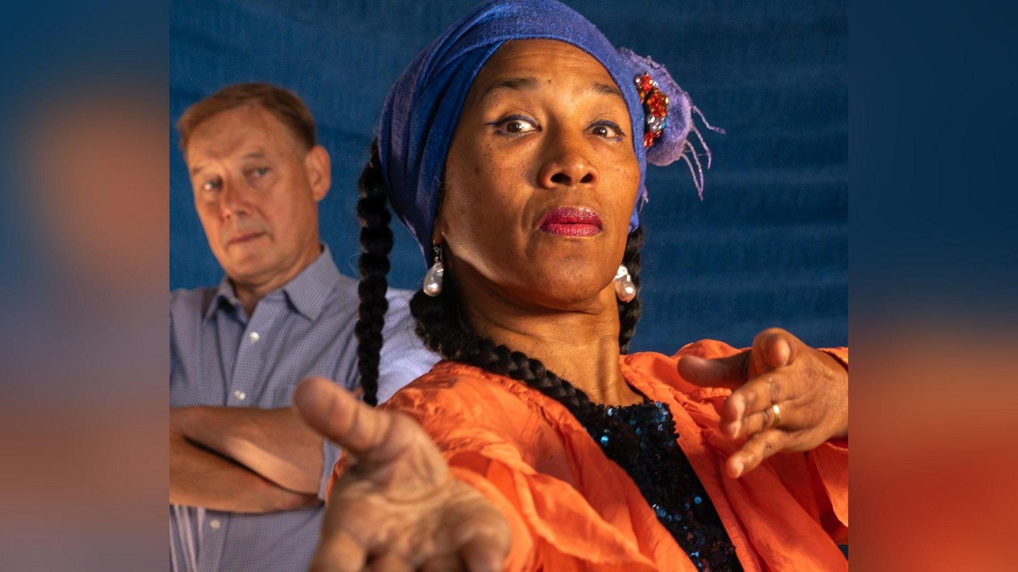 Mary-Ann Roberts yn gwisgo siaced oren a chlustlysau perlog. Mae Richard Parry yn y cefndir yn edrych arni ac yn gwisgo crys glas golau.