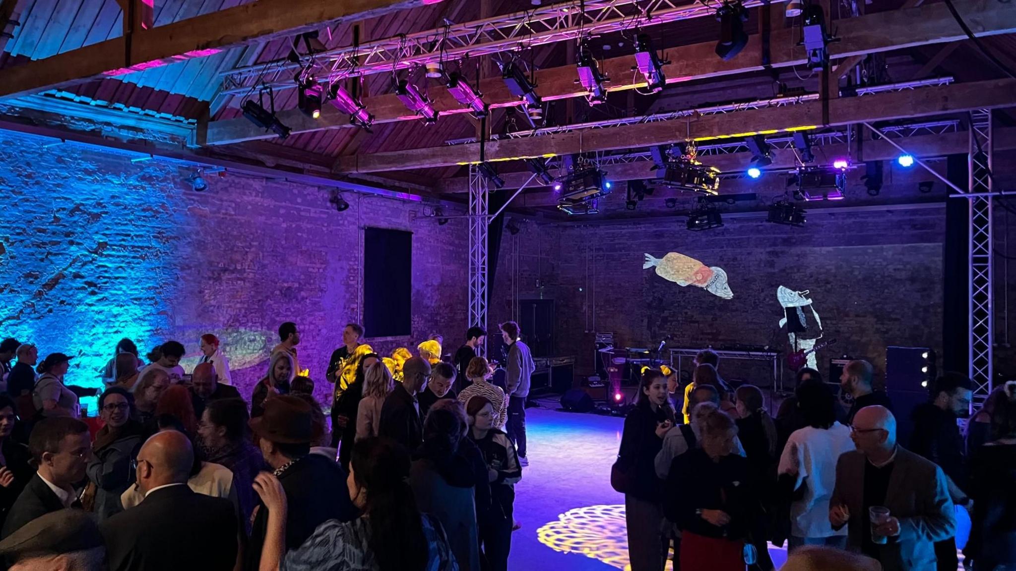 The interior of Great Yarmouth's Ice House building. Brickwork is bathed in blue and purple light. There are people milling around at a launch event. A stage area has been created at one end with artworks of two fish on that wall.