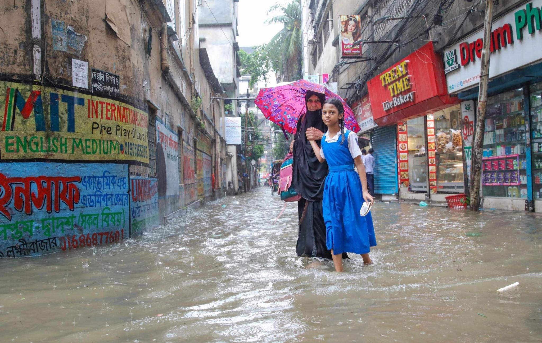 A schoolgirl in a blue and white pinafore and a woman in a black dress carrying a pink umbrella wade through ankle high waters in a flooded street in Dhaka