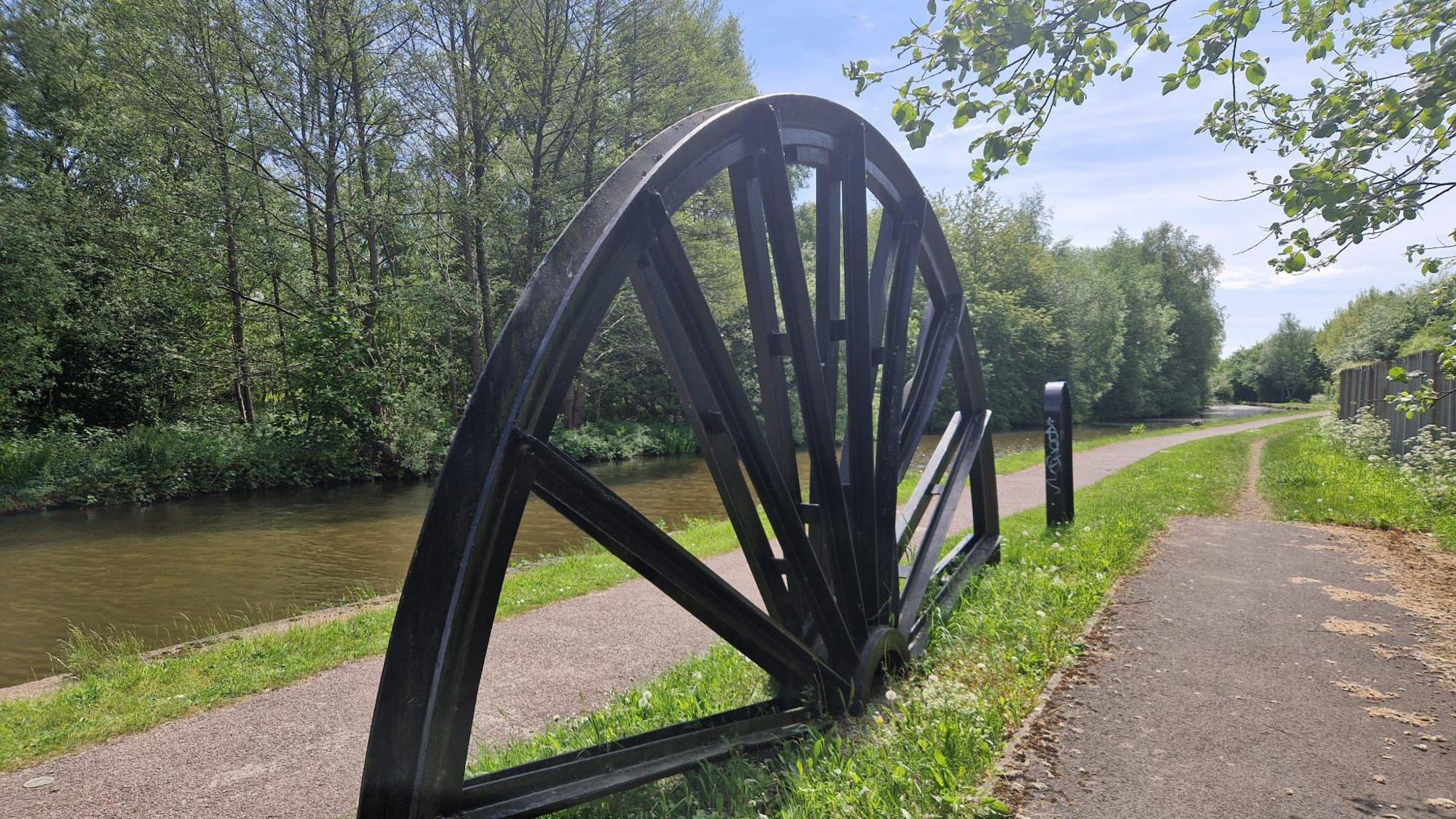 A canal is in the far left of the image and runs along the top. There are two paths one of which is the canal towpath and they are separated by a black landmark, which is half a wheel on a thin stretch of grass. The sky is pale blue with a few clouds and a tree drapes into the right-hand corner of the image. 