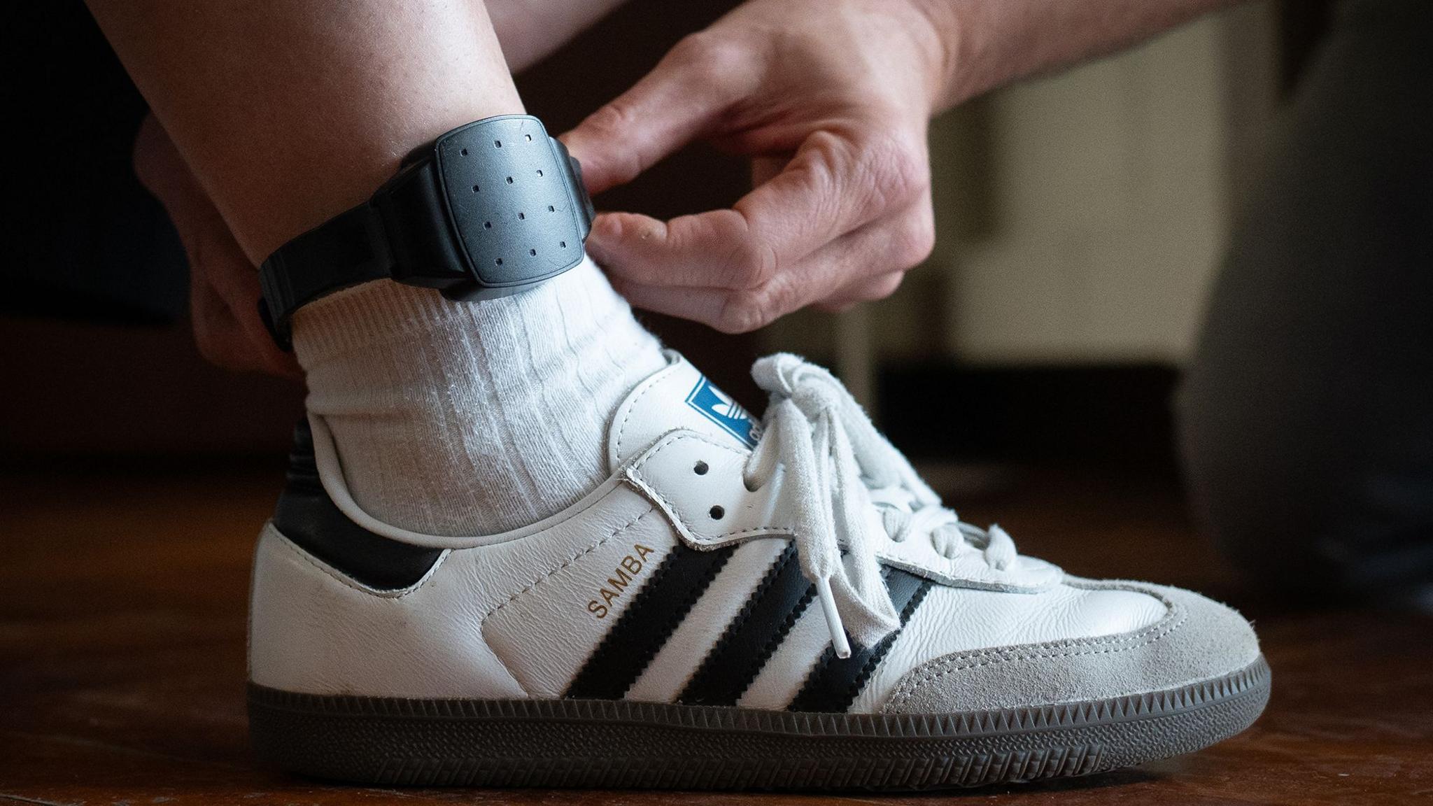 A lightweight tracking device is fitted to a man's ankle. The photo is a close-up of a trainer, white sock and lower part of the man's leg