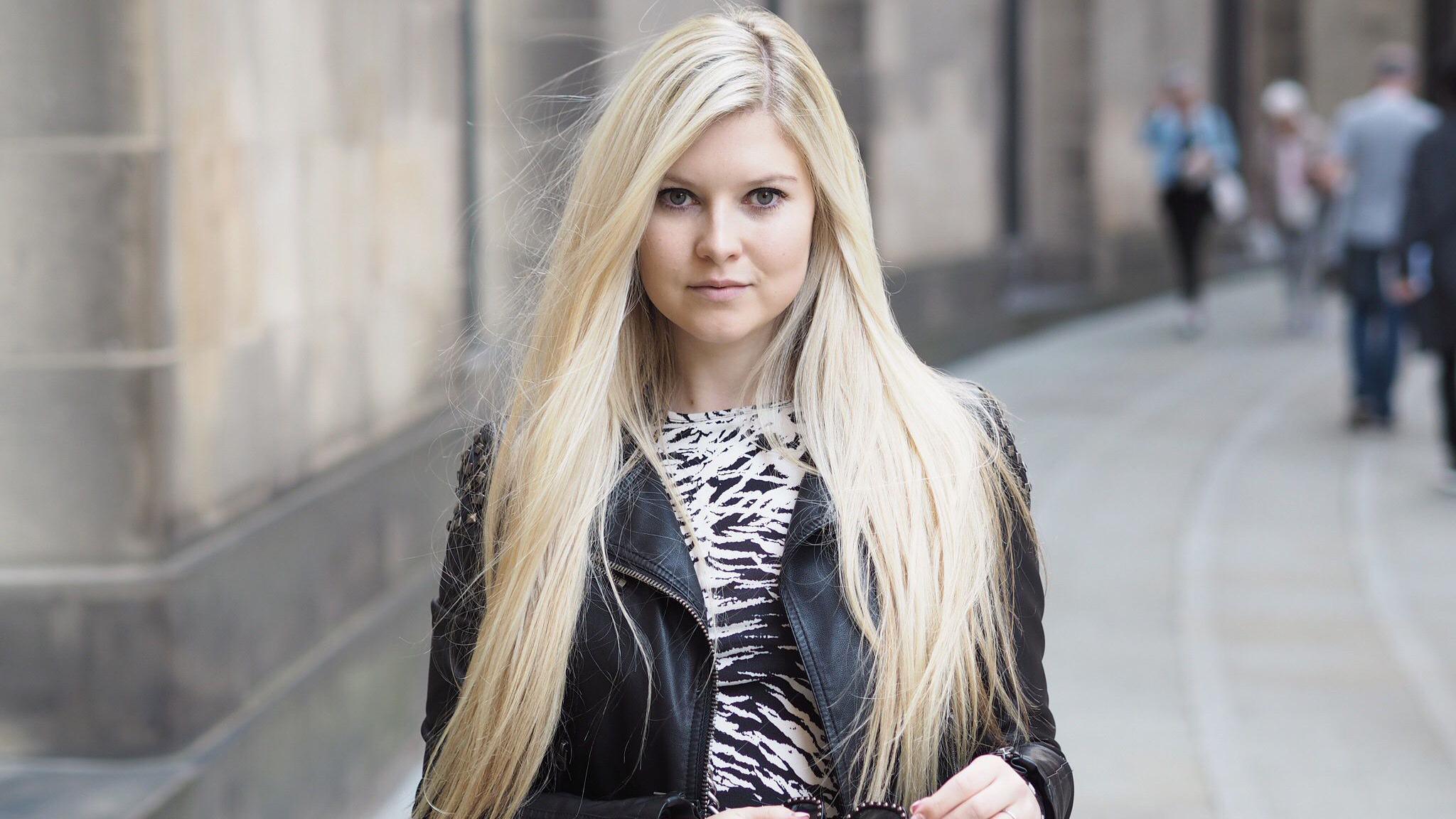 A woman with long blonde hair, wearing a black leather jacket and white and black striped top, stands in a street while holding a pair of sunglasses.