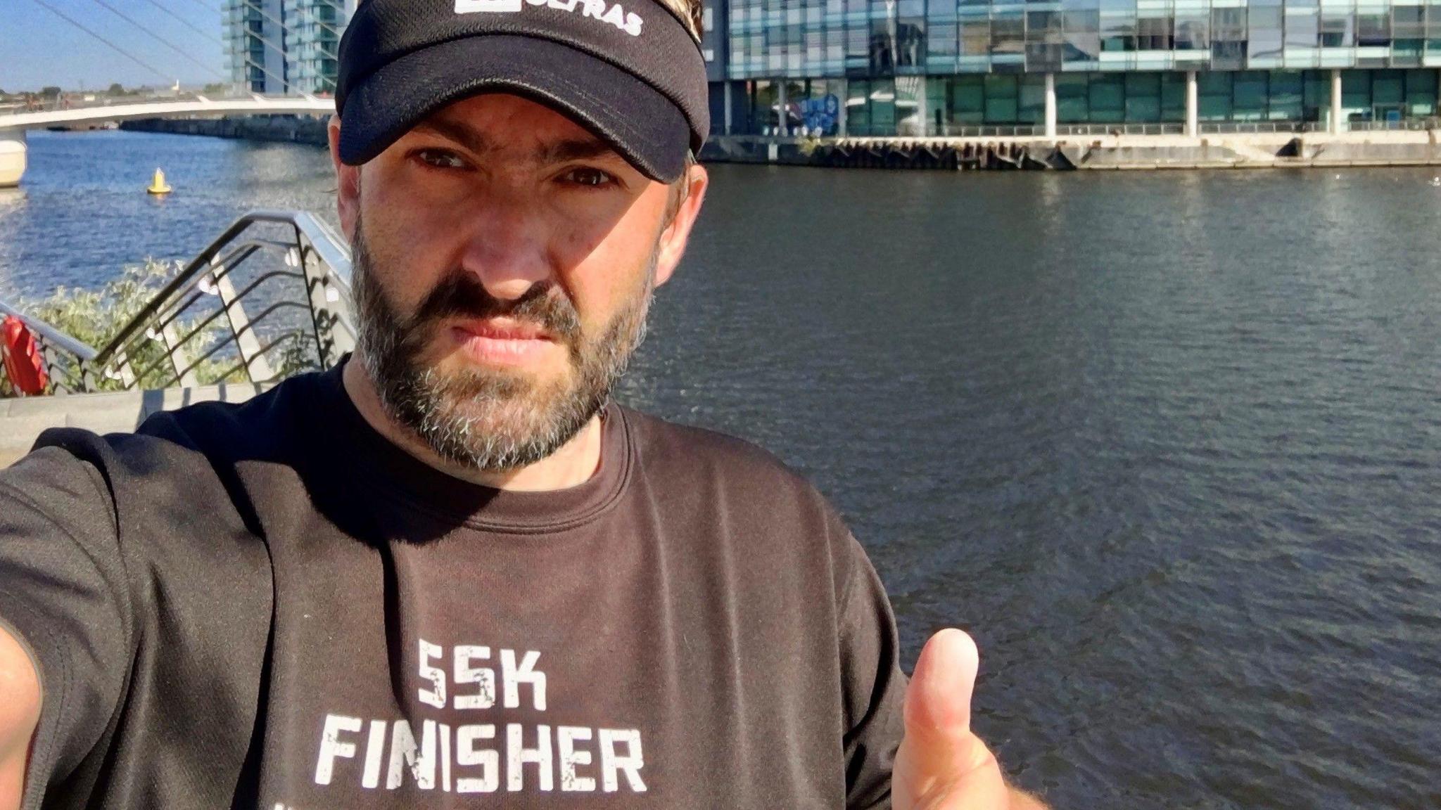 A man takes a selfie at Salford Quays. There is a glass building, a bridge a large canal in the background. He is wearing a hat, has a greying black beard and is wearing wearing a black tshirt that says '55k finisher'