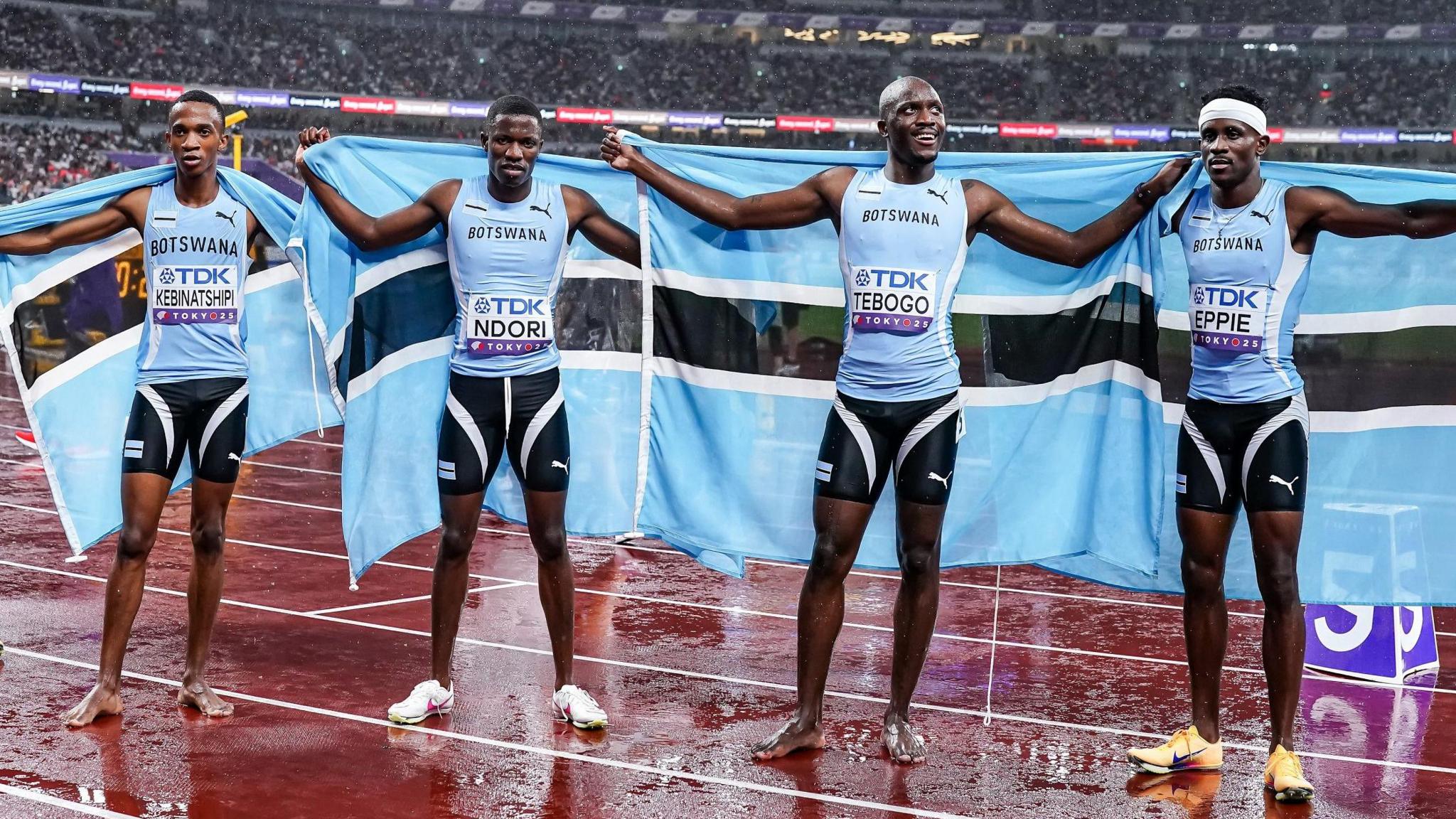 Collen Kebinatshipi, Bayapo Ndori, Letsile Tebogo and Lee Bhekempilo Eppi, all wearing blue running vests and black shorts, each hold a Botswana flag behind them while standing on a wet running track. Kebinatshipi and Tebogo are both barefoot while Eppi wears a white headband. Stands of a packed stadium are seen in the distance behind them