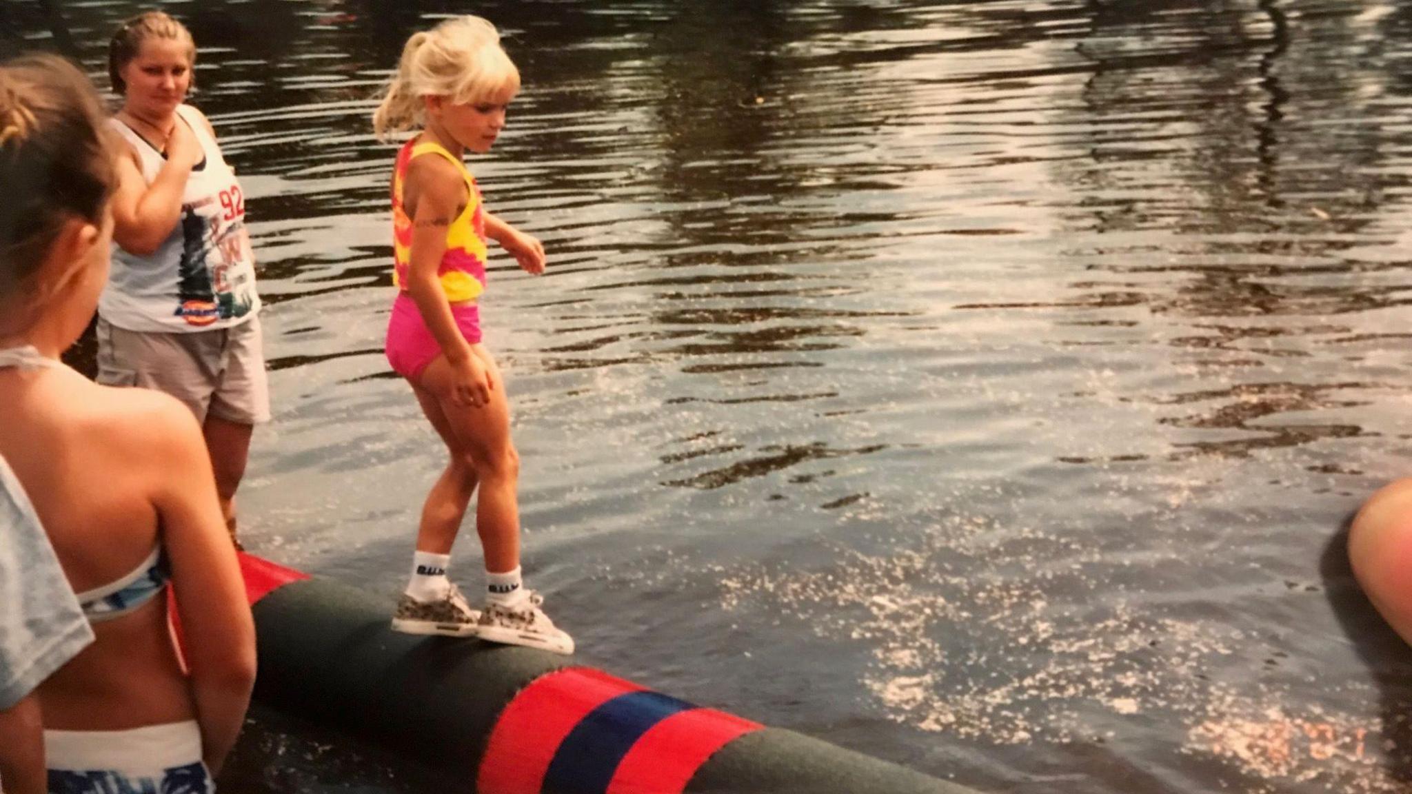 A blond child stands on a log in the water, balancing in socks, sneakers, pink shorts and a pink and yellow top, her blonde hair tied up in a ponytail