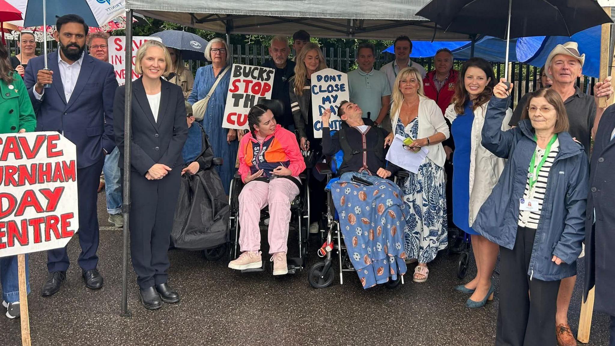 A line of people standing under a gazebo. Two of them are in wheelchairs. Some are holding umbrellas. Some are carrying placards which say "Bucks stop the cuts" and "Don't close my day centre".