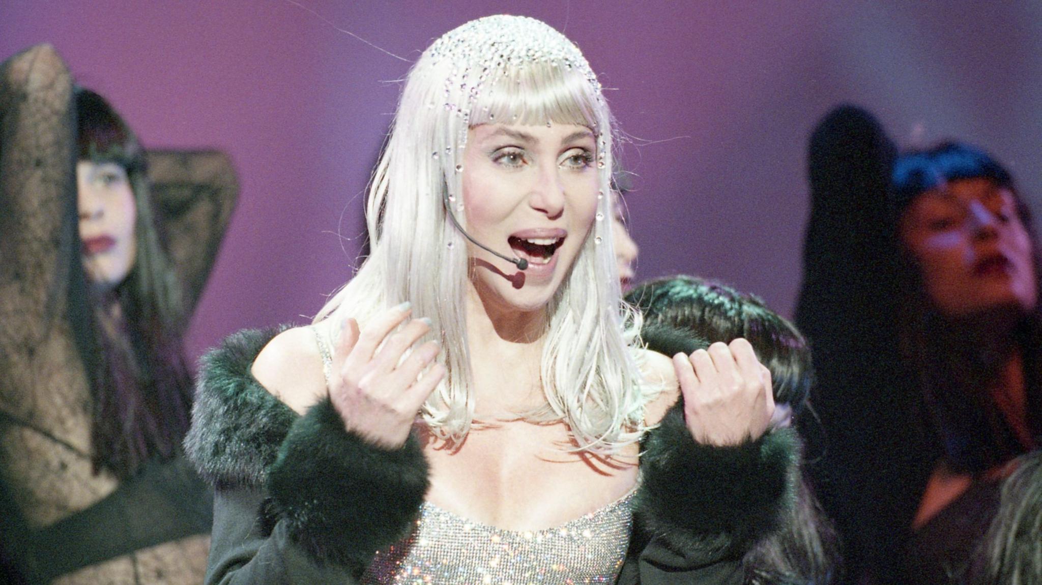 Cher performing in the the 1990s wearing a microphone headset and wearing a sparkling silver dress beneath a black top. Two dancers are in the background