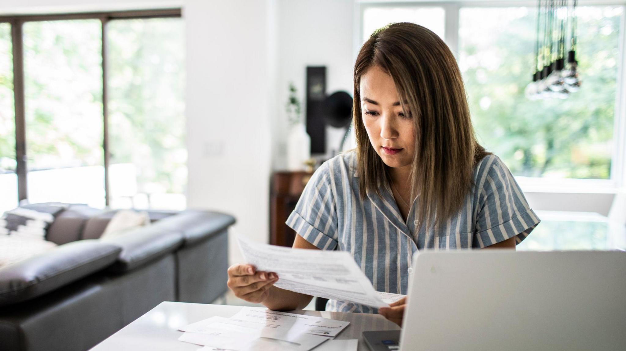 Woman wearing striped shirt looking at bills while sitting at laptop in her home