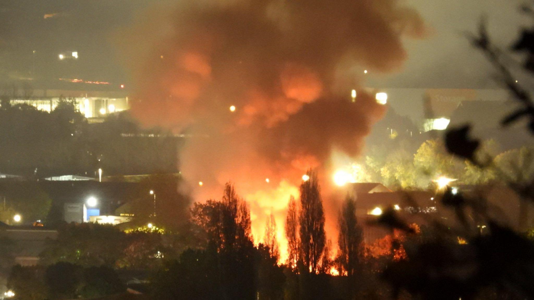 A large plume of smoke in the night sky at a distance. There are trees in the foreground. There is an orange glow from the flame at the bottom of the smoke. There are some buildings surrounding the blaze