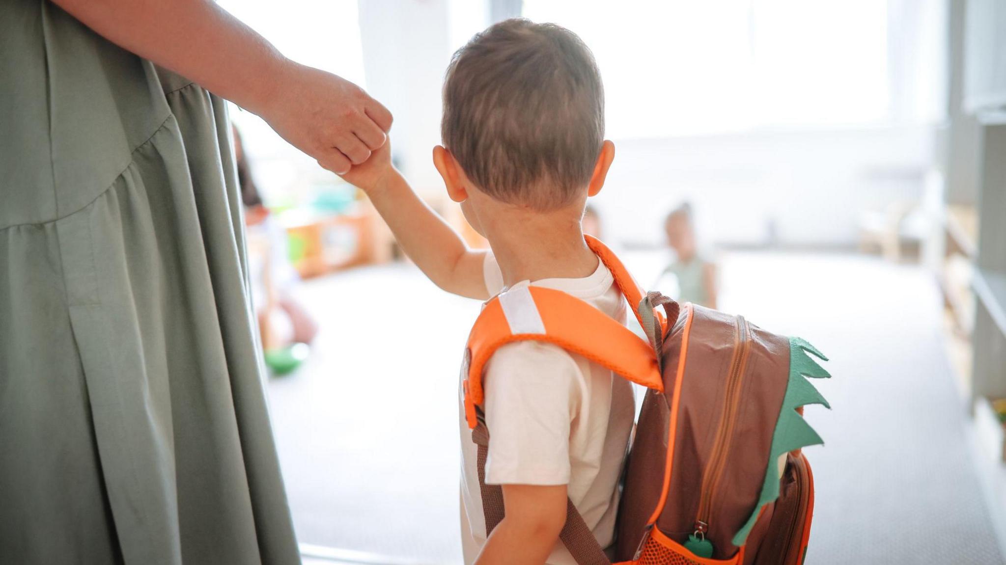 A toddler with an orange and brown backpack holds a woman's hand, a day care centre in the background 