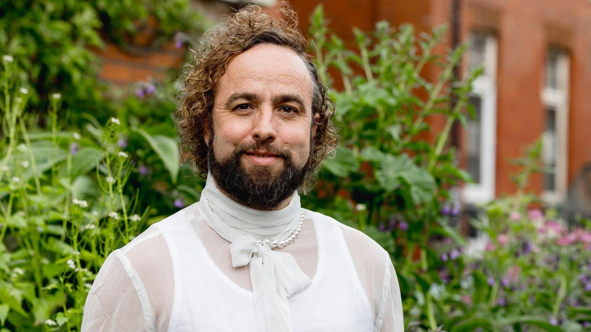 Mae Daf James o flaen gardd ac yn gwisgo crys gwyn. Mae ganddo wallt brown cyrliog gyda barf. Mae e'n wenu. 