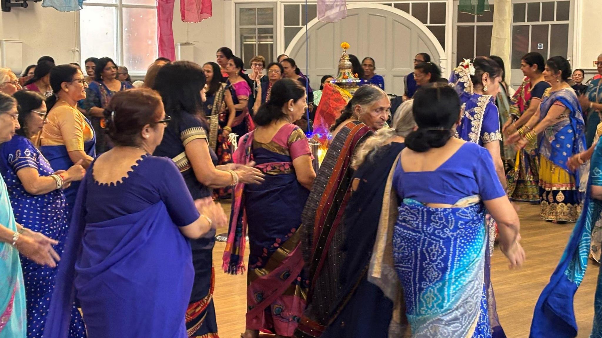 A group of Indian women in colourful vibrant sarees, dancing the energetic Giddha with its clapping and call-and-response singing. This circular dancing is often performed during Navratri celebrations and formerly performed by women in temples focusing on religious stories. The devotions have an alter at the middle of the space.