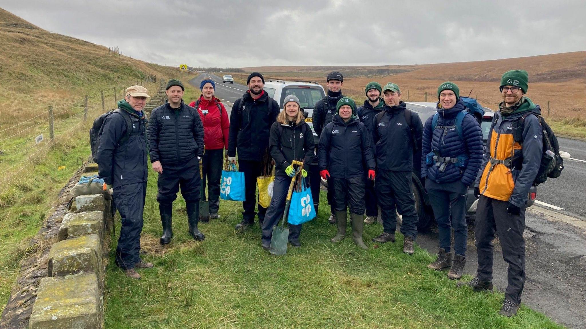 Eleven men and women in outdoor, waterproof clothing standing in a layby next to moorland in West Yorkshire. Some are carrying carrier bags full of saplings, others are holding spades.