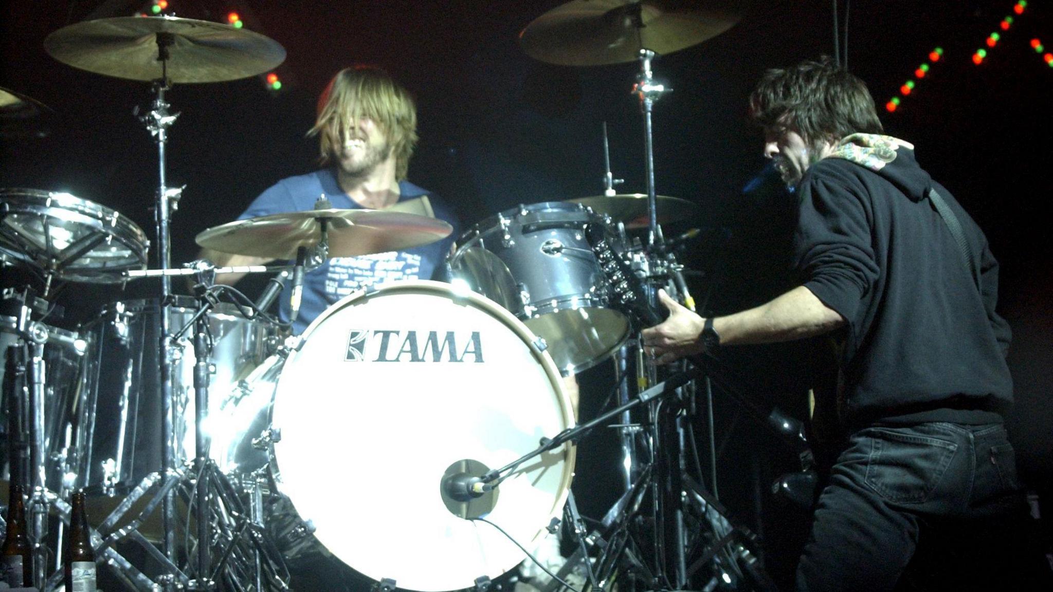 Taylor Hawkins, who has long blond hair and a beard, plays drums while Dave Grohl, dressed all in black with black hair and a beard, plays guitar facing him