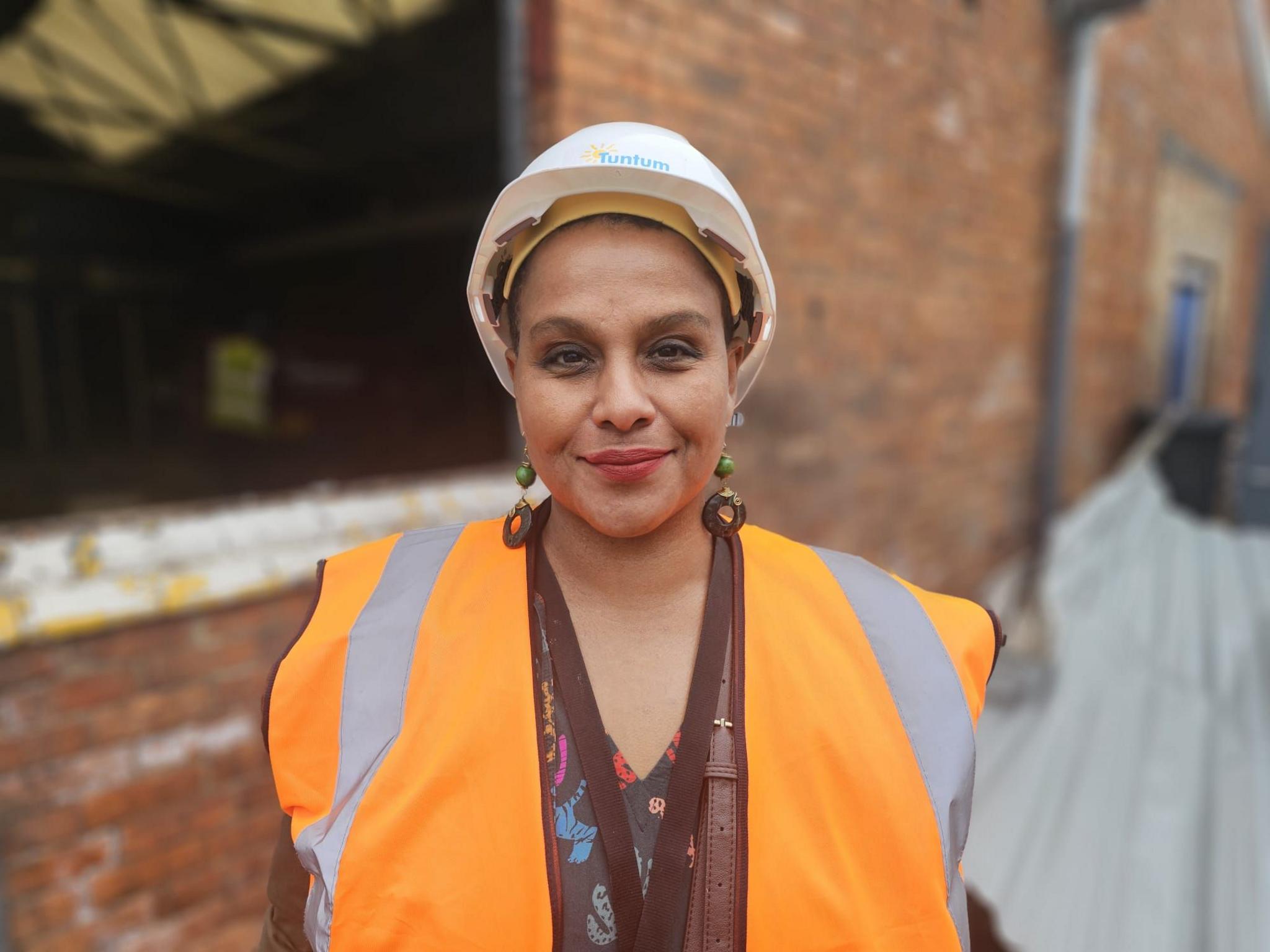 A woman wearing a white hard hat and orange high-vis vest. She has large earrings and a brown bag strap over her shoulder.
