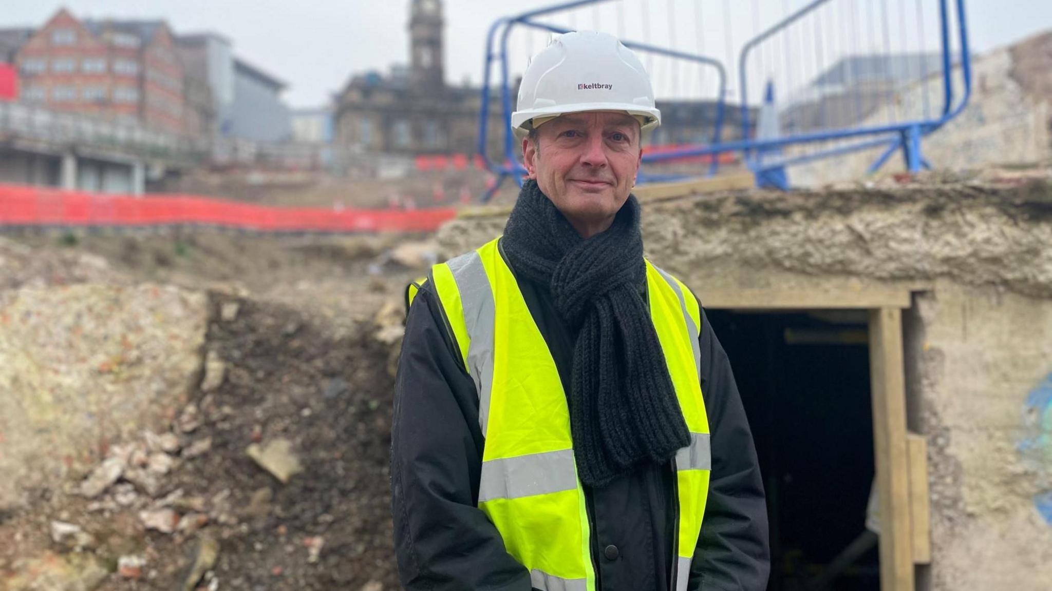 Martin Gorman, wearing a hard hat and bright yellow jacket, stands in front of stone remains and an open chamber