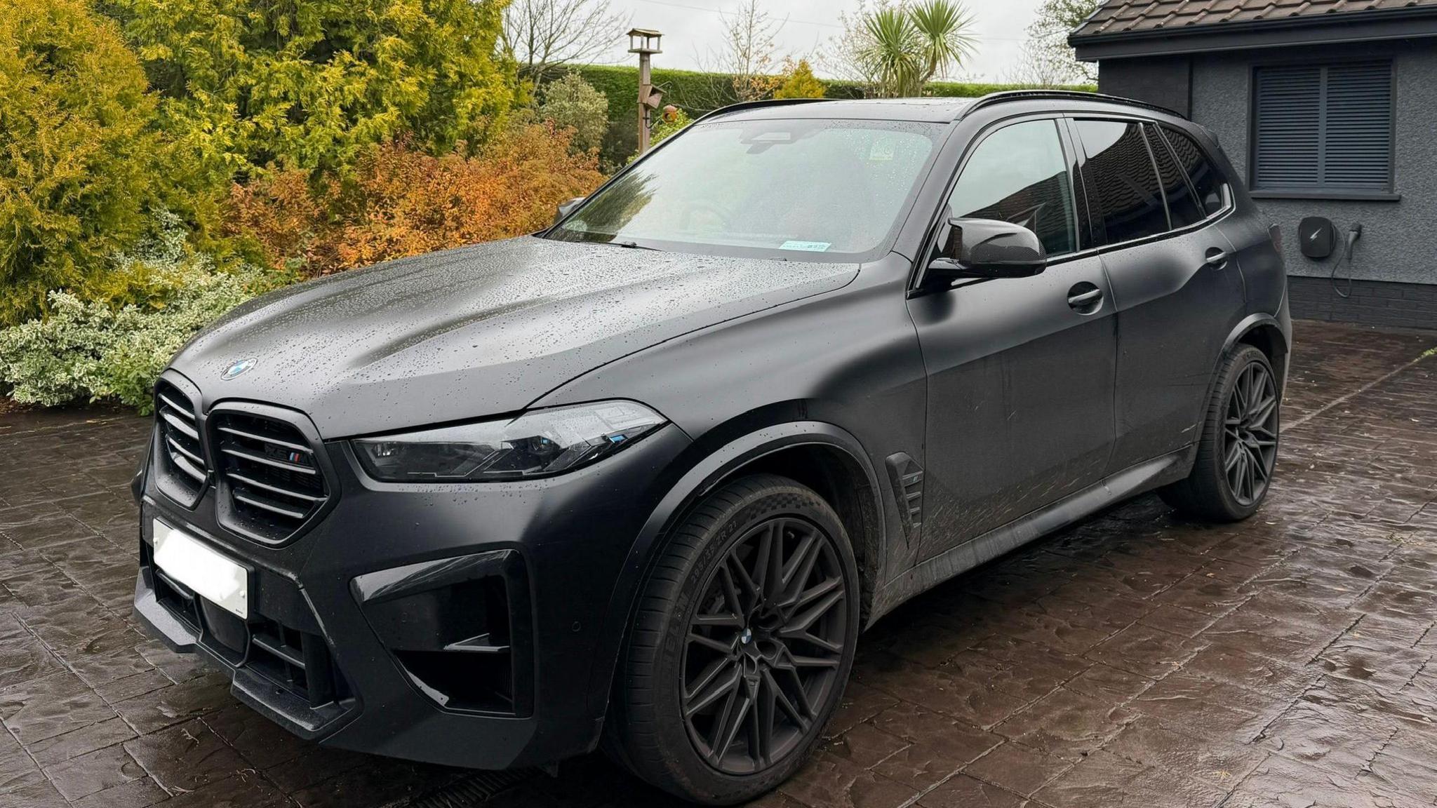 A large matte black BMW jeep sitting in the rain on a paved driveway with a garden visible behind it.