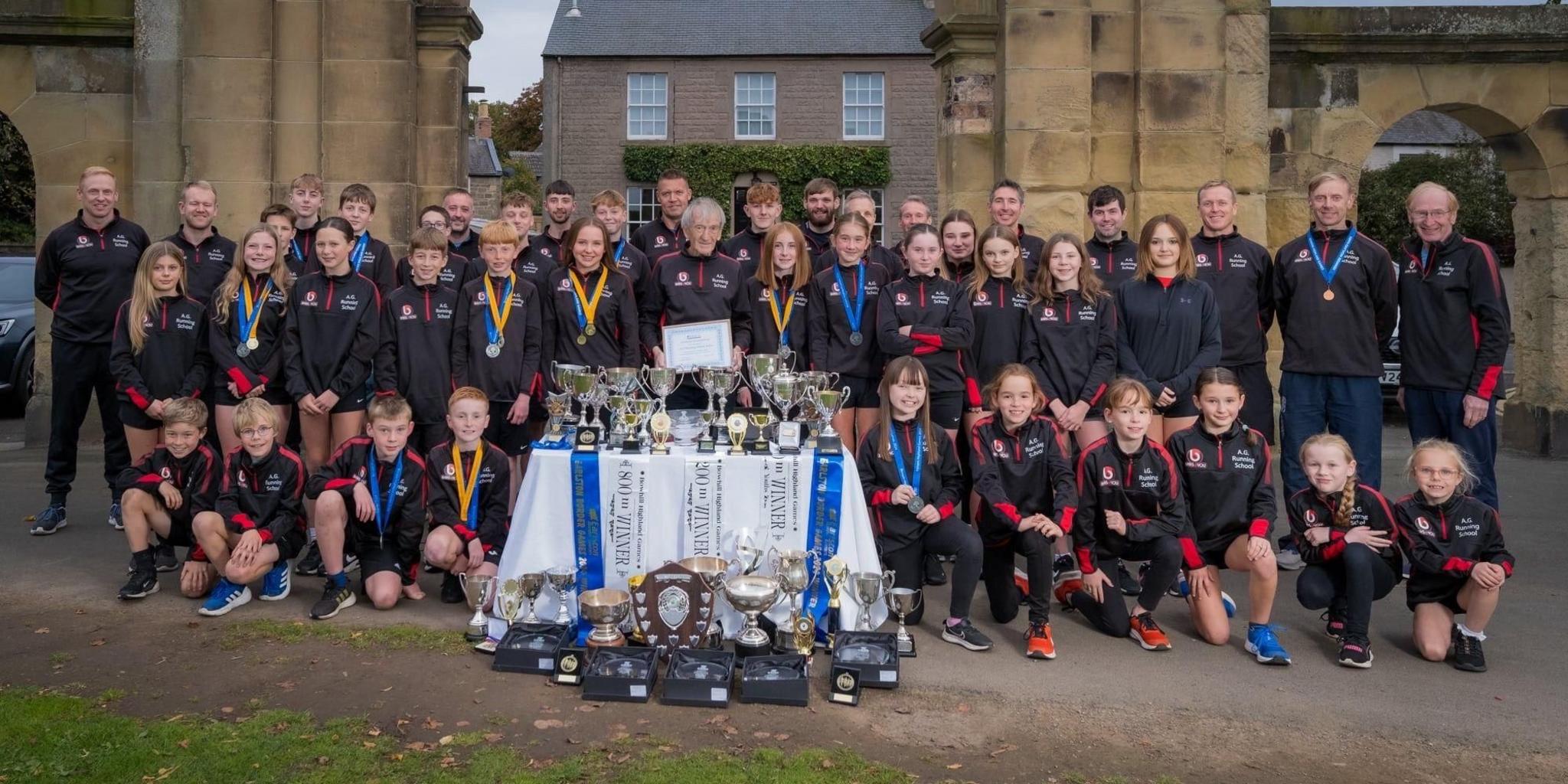 A group of three rows of athletes in navy tracksuits, some with medals around their necks, flank coach Addie Gray in centre, behind a table bedecked in trophies, at the entrance to Kelso's Shedden Park.