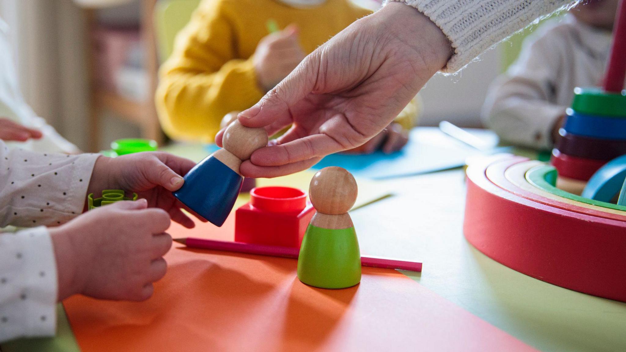 Colourful toys on a nursery table, one of is being held by a child and an adult simultaneously. We can only see their hands and sleeves.