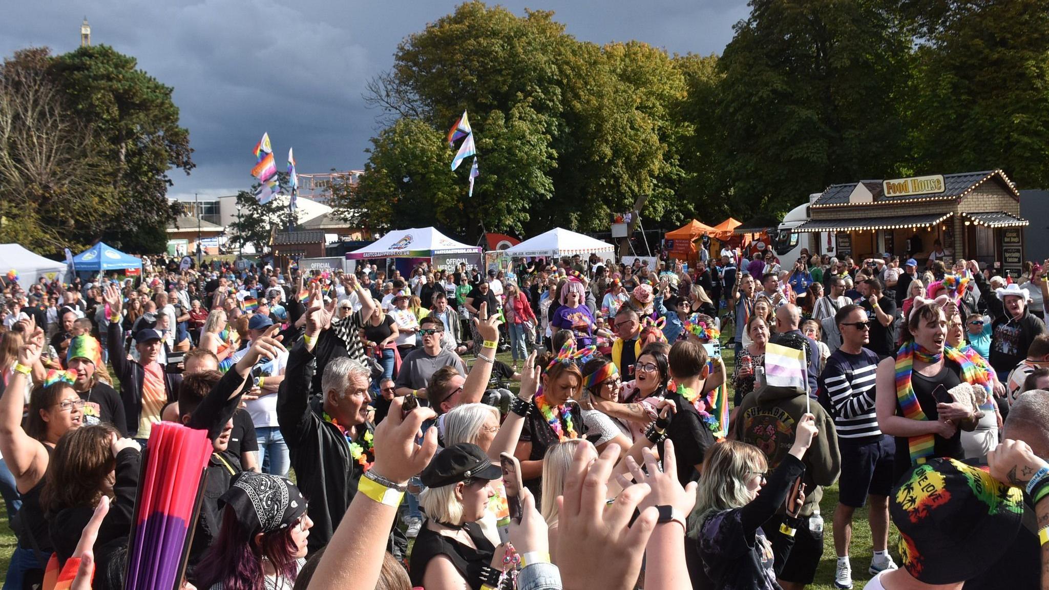 A picture of the crowd in the sunshine by the main stage. Lots of people have their hands in the air.  Some festival flags are flying in the background.