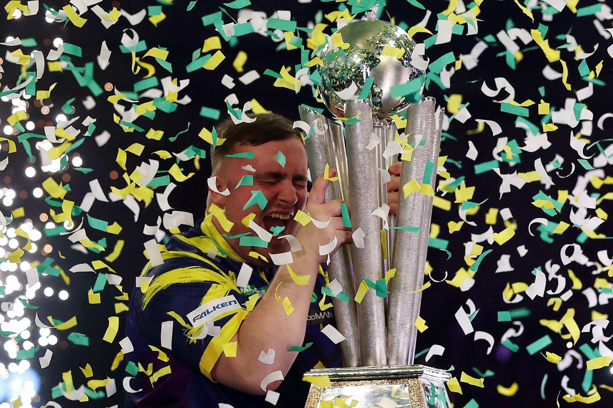 A person wearing a dark blue and yellow jersey holds a large silver trophy with multiple vertical columns and a globe-like top. Green, yellow, and white confetti is falling all around.