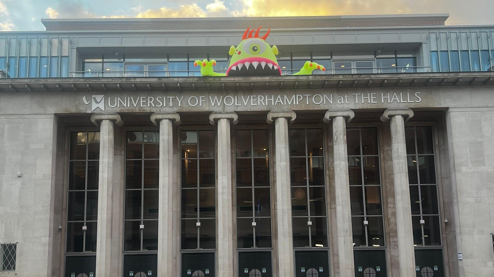 A green inflatable monster with one huge eyes and sharp teeth looming over The Halls concert building in Wolverhampton. The building is grand and has six huge columns at its exterior. 