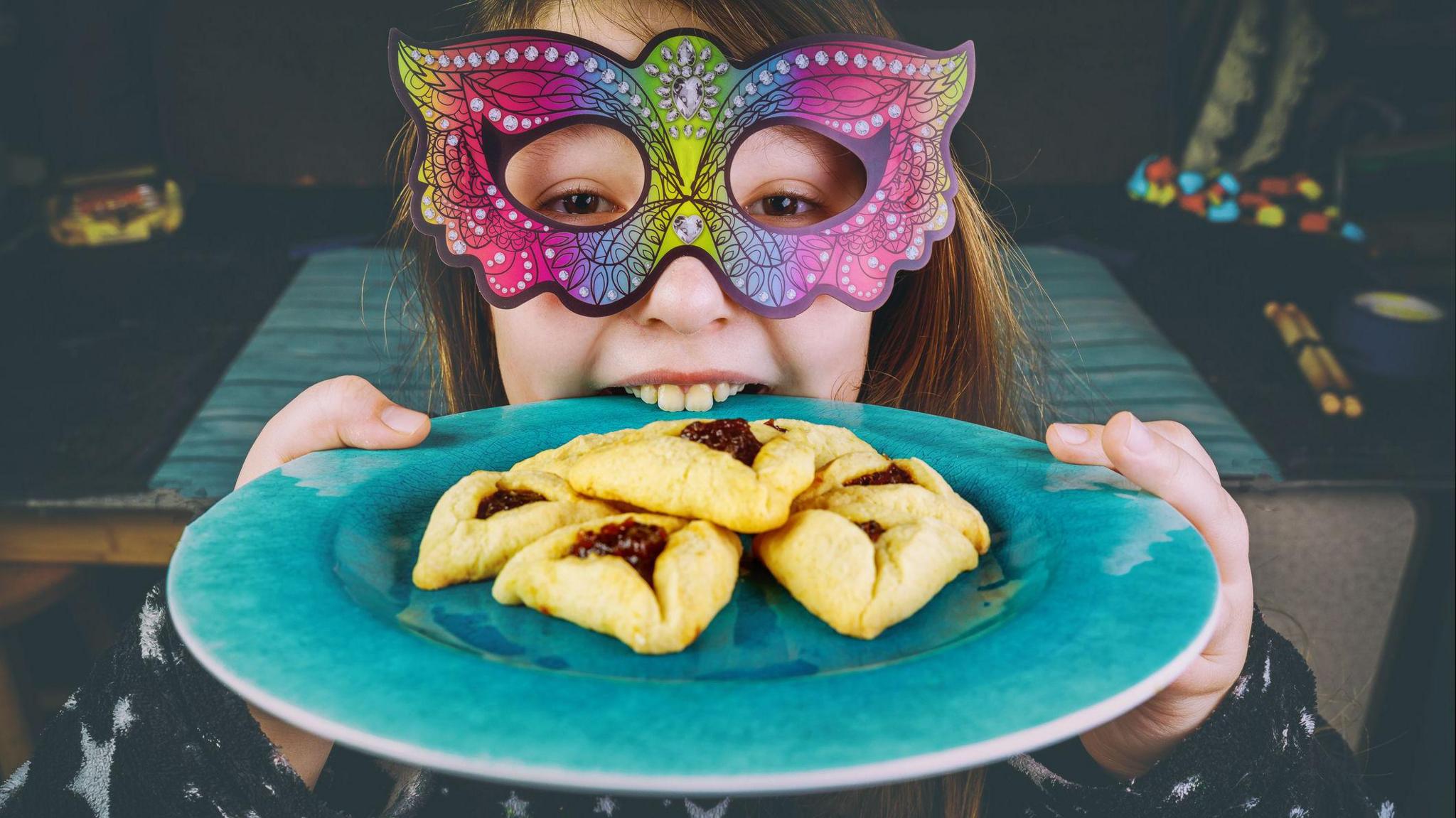 A girl wearing a colourful mask holds a plate containing triangular-shaped sweet pastries called Hamantaschen. 