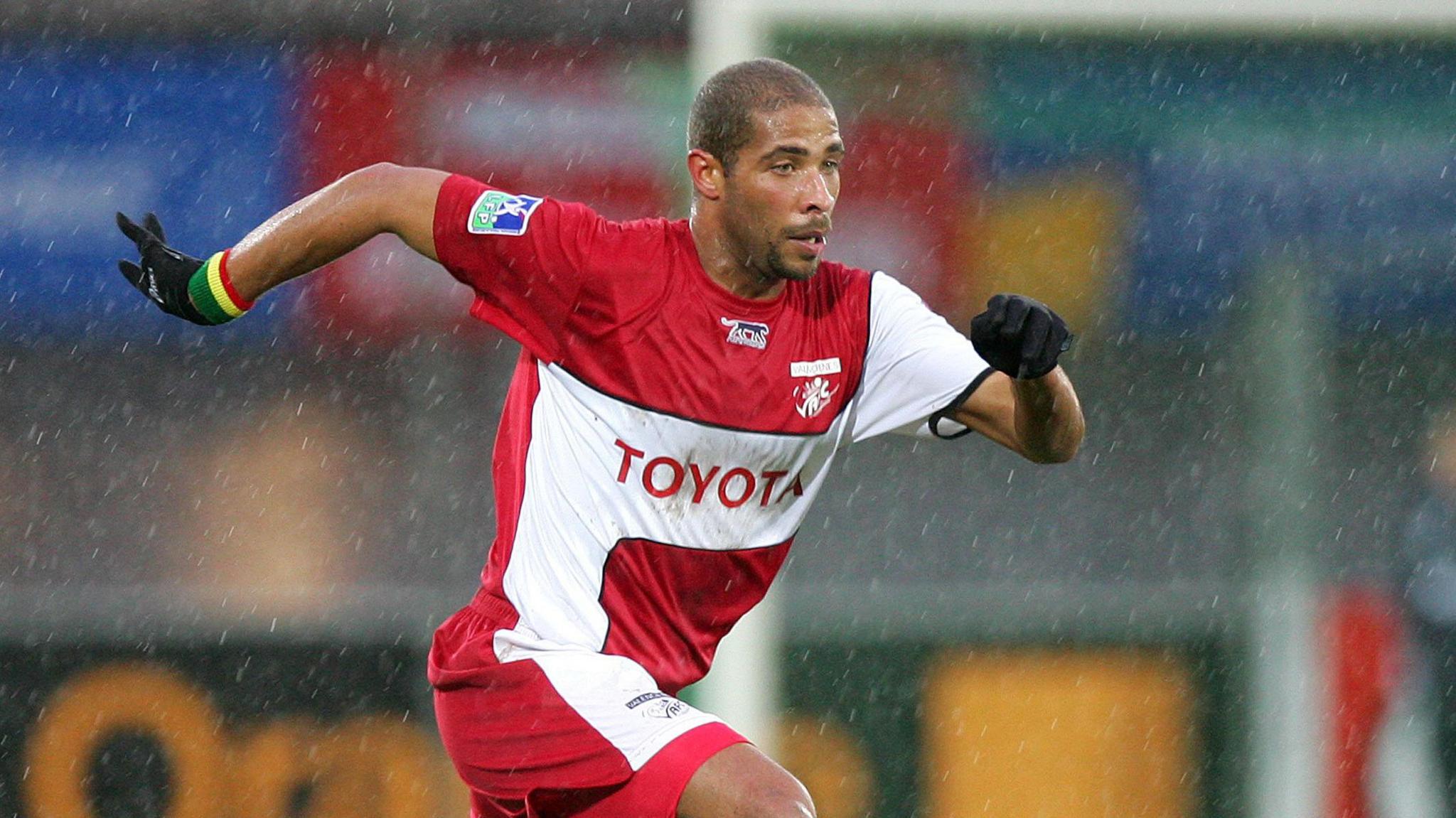 Eric Chelle, wearing a red and white Valenciennes shirt, black gloves and a sweatband in green, yellow and red on his right wrist, runs during a football match on a rainy day