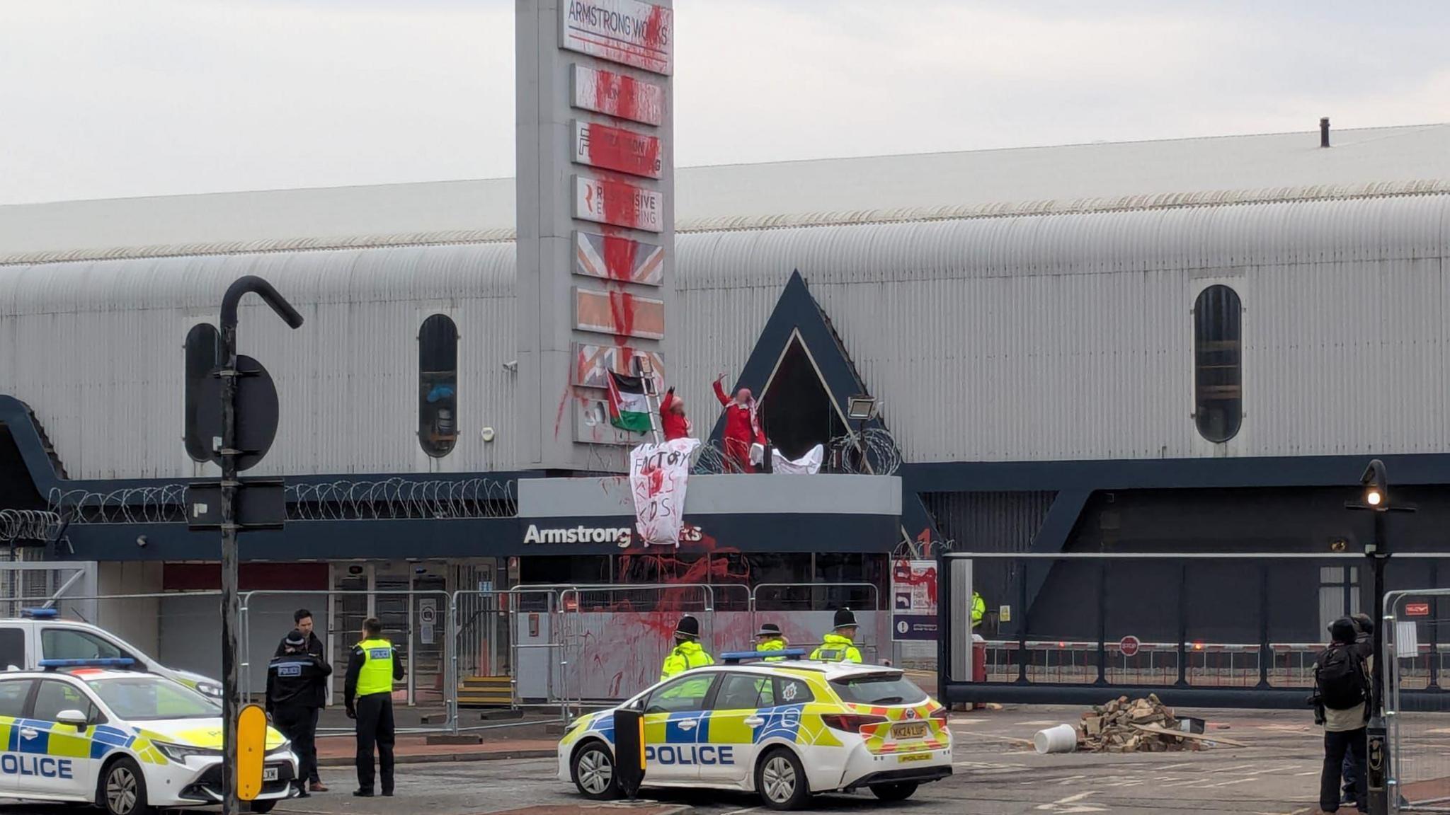 Two female protesters wearing red jumpsuits on the gatehouse of the Armstrong Works building. Red paint has been splashed up the factory's signs. A pile of rubble has been placed in front of the gate. Three police cars and several police officers stand outside the factory.