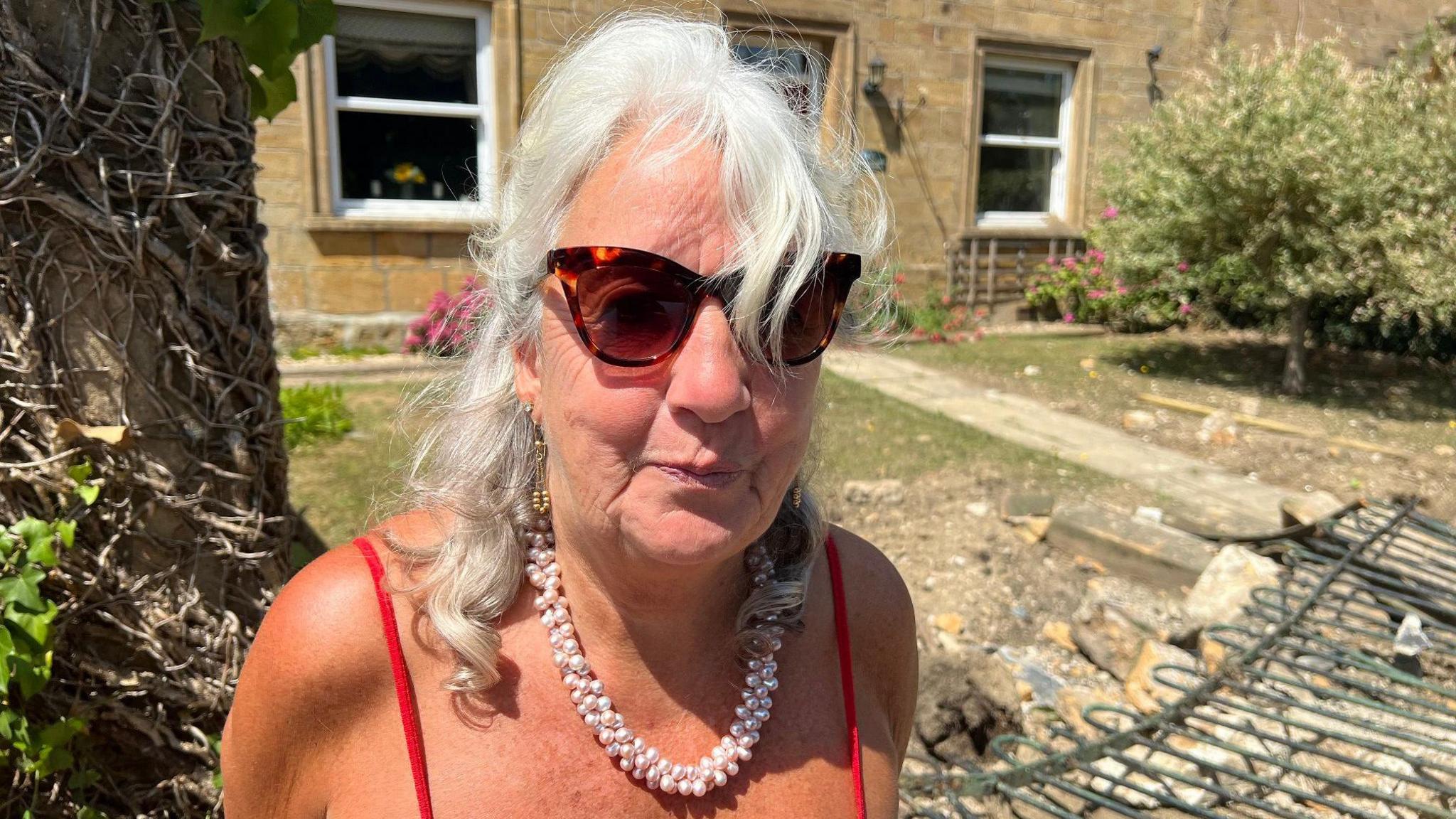Claire, who wears a red strappy top, a large pearl necklace and big sunglasses, smiles at the camera. She is standing in front of the fence, which has been knocked down, and rubble can be seen strewn across the garden.