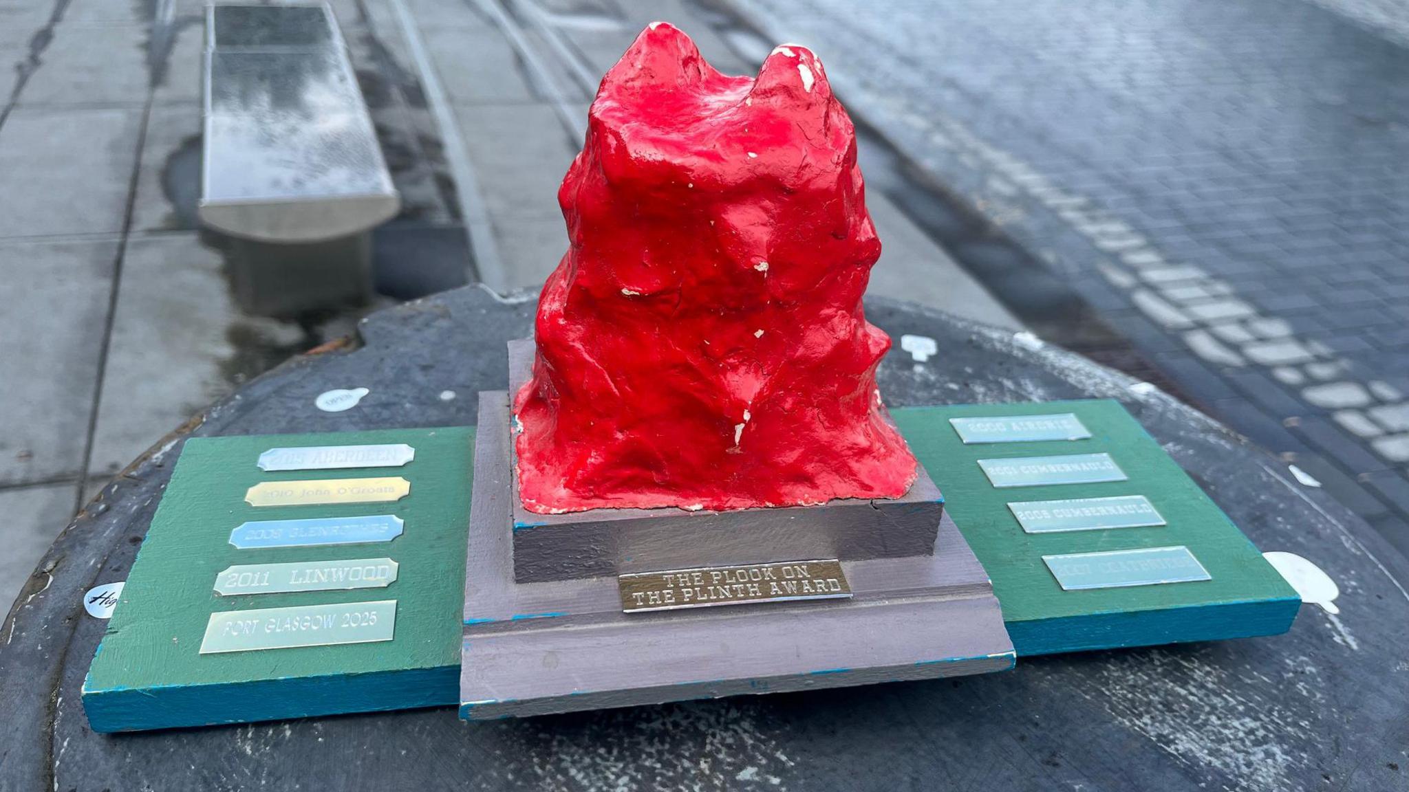The award is a red textured object on a wooden plank. It features engraved writing which reads "THE PLOOK ON THE PLINTH AWARD". Other plaques on the award feature the names of towns who previously won the award, like Linton in 2011. The award is on top of a bin on a street.