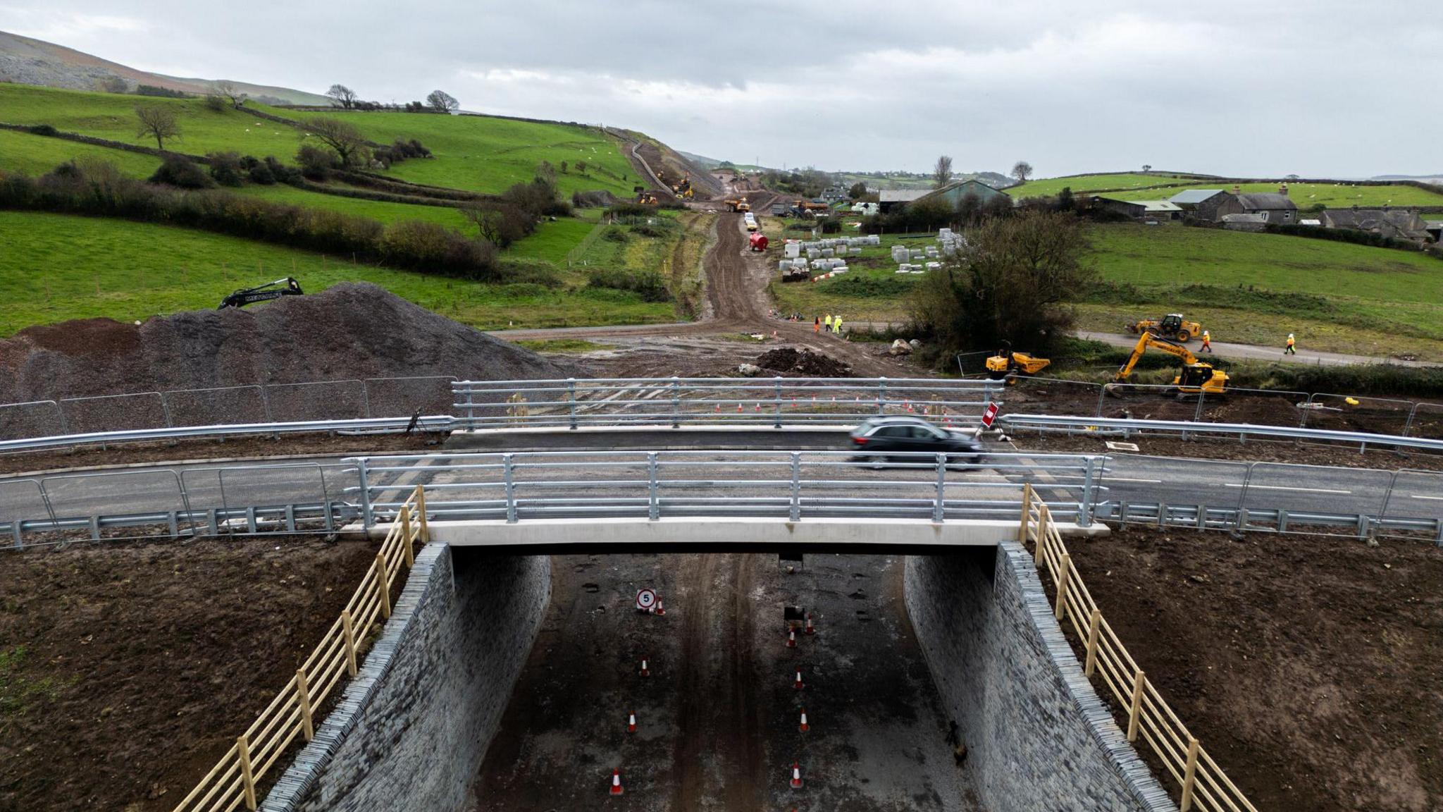The new bridge with a car in motion travelling on it. The bridge is over an unfinished stretch of road, which looks like a dirt track, with machinery and workers in high viz working on the site.