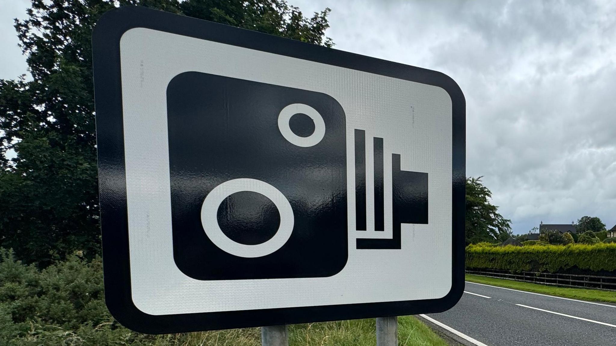 A white road sign with a black speed camera symbol. It has a black border. Behind on the right is a road with green hedges above. To the left is green grass and trees.