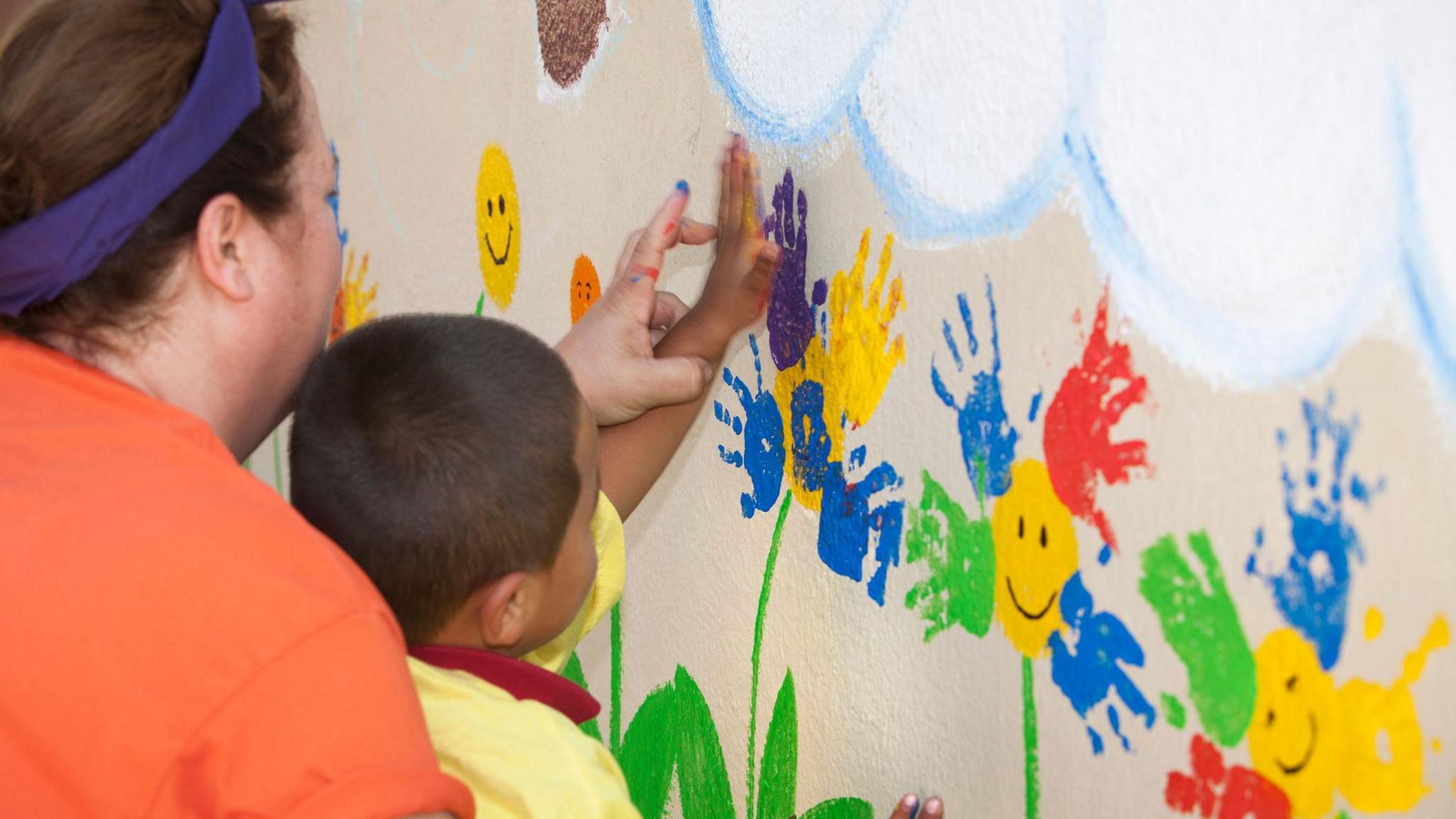Woman helps young child hand paint on a wall 
