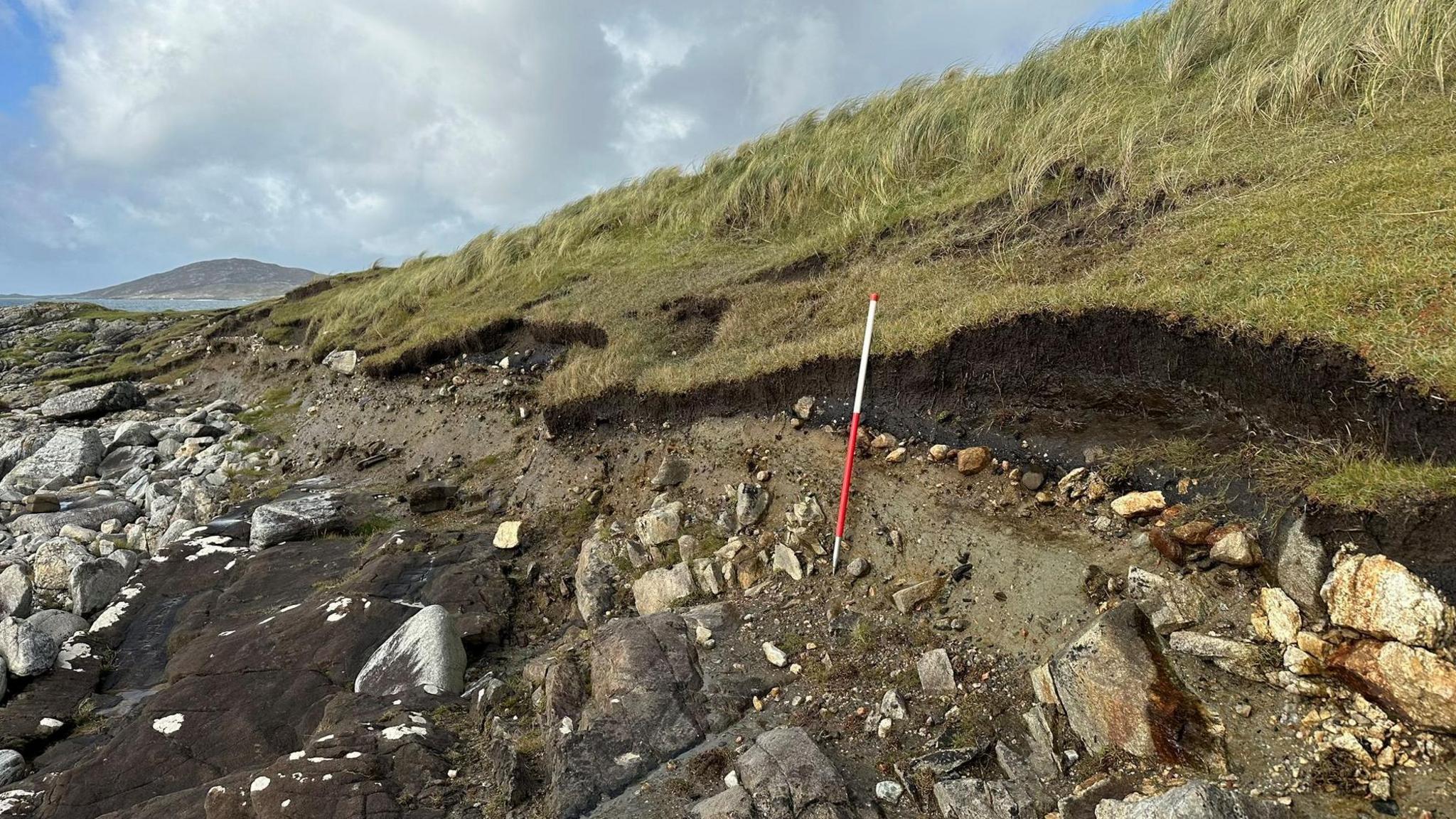 An area of shorelines has been washed away to reveal a charcoal-rich layer. There are rocks below a grassy bank.