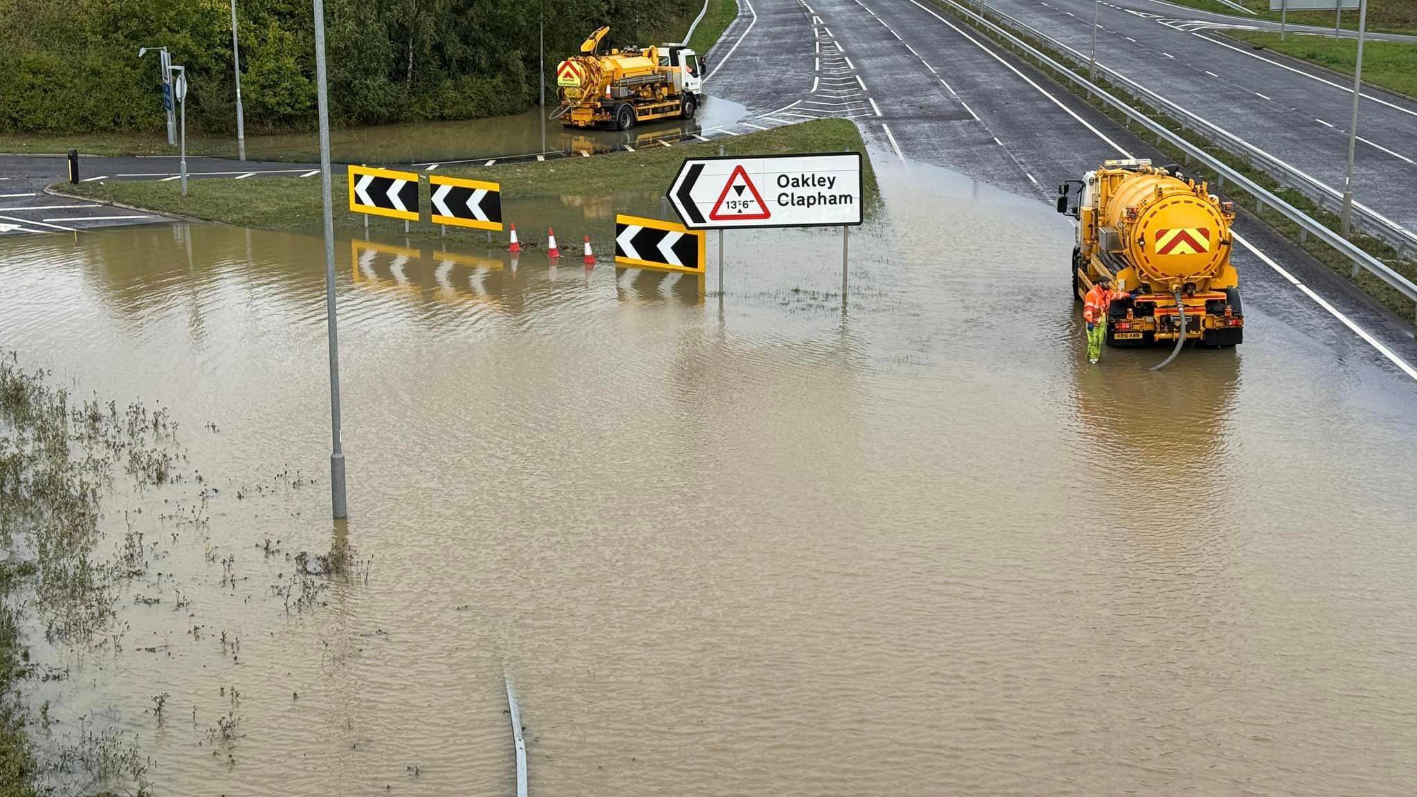 Yellow tank vehicles pump water out of a flooded road junction with a sign saying Oakley Clapham