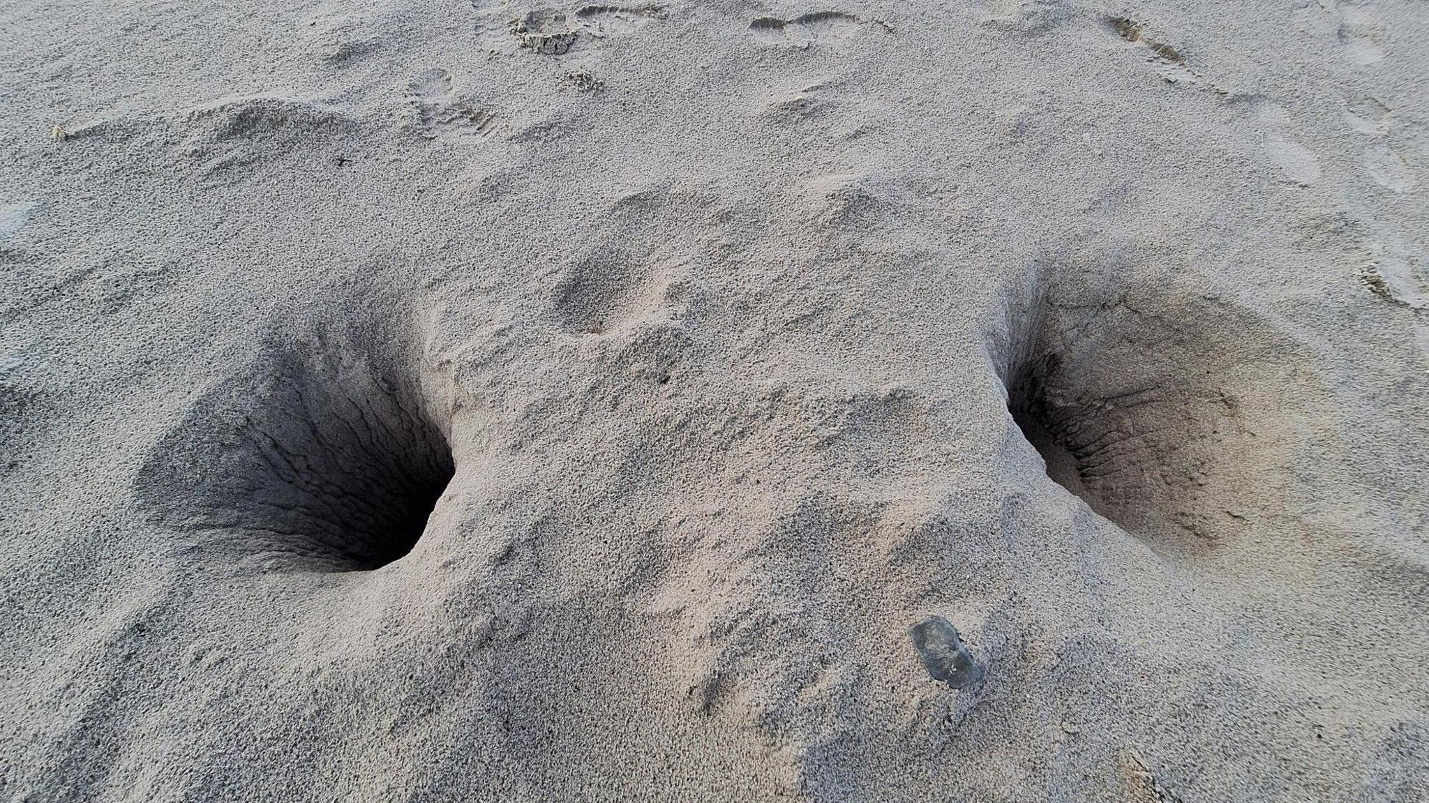 Two large holes on a sandy beach. Both holes have signs of scratches in the sand on the sides of them. There are footprints in the sand at the top left of the picture.