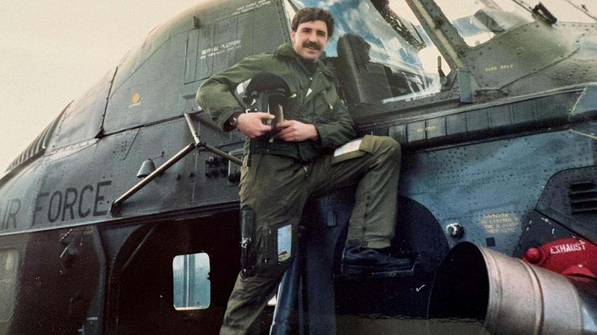 A man is standing on the side of a helicopter. He is dressed in uniform and is holding a helmet. He has dark hair and a moustache and is smiling at the camera.