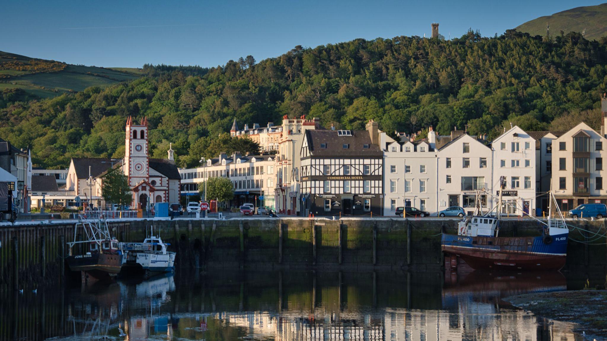 Ramsey quayside overlooking the harbour, buildings like the quay in front of a leafy hill with the Albert Tower above, on a sunny day.