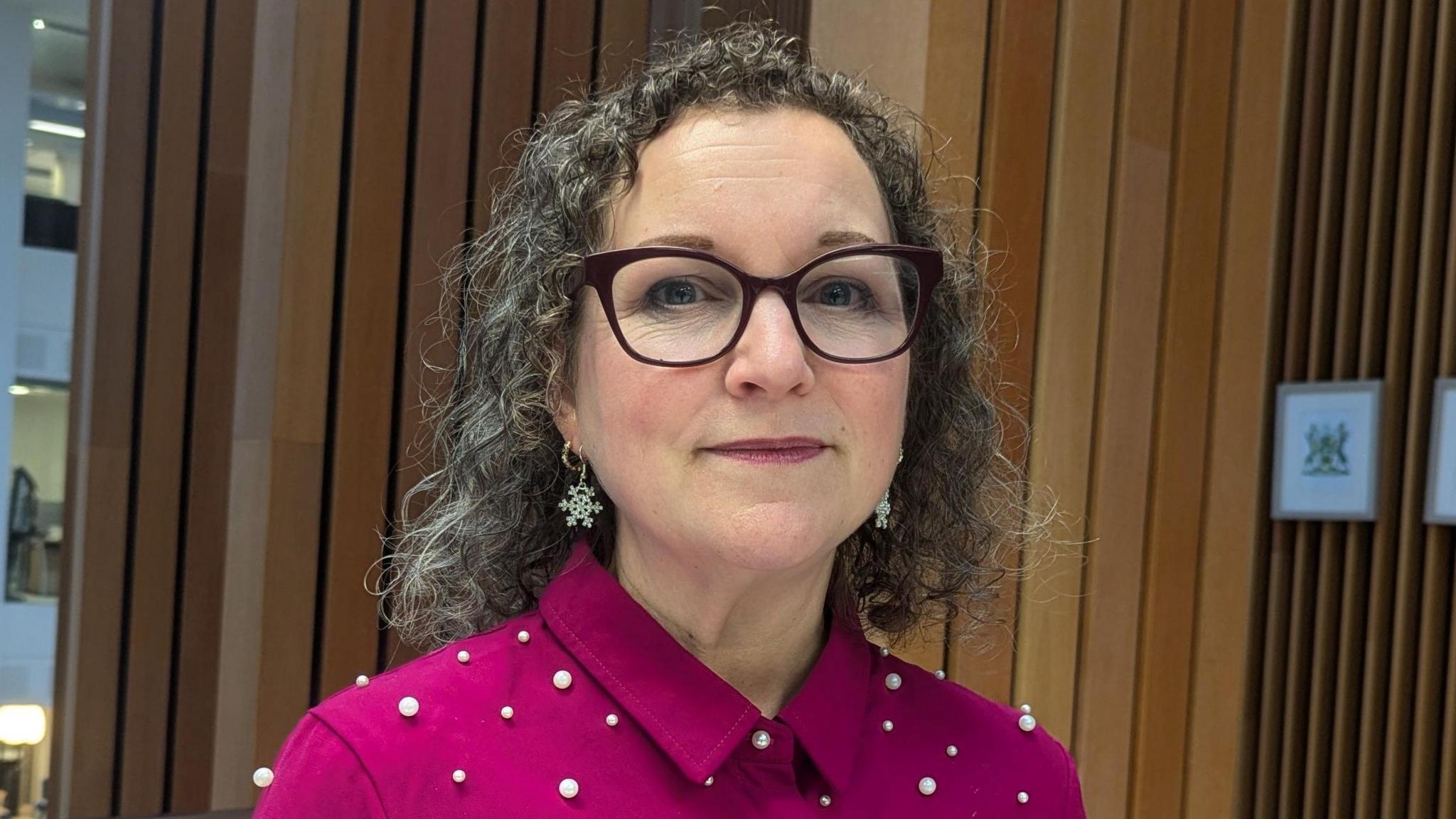 Image of Nadine. She is wearing a pink shirt with pearl-like object on. She has glasses on and has shoulder-length hair.