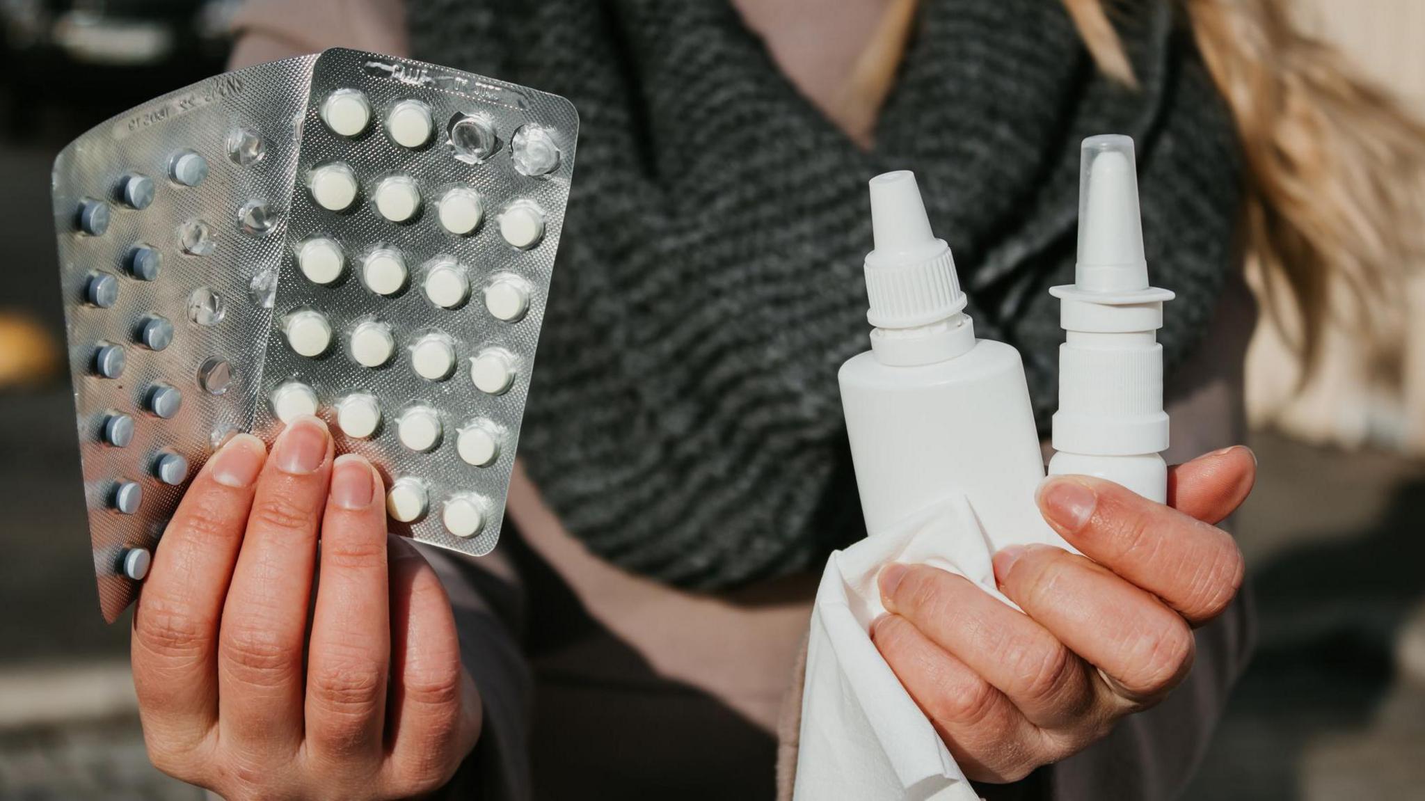 A woman holds up two packs of antihistamine pills in one hand and nasal and eye sprays on in the other, along with a tissue