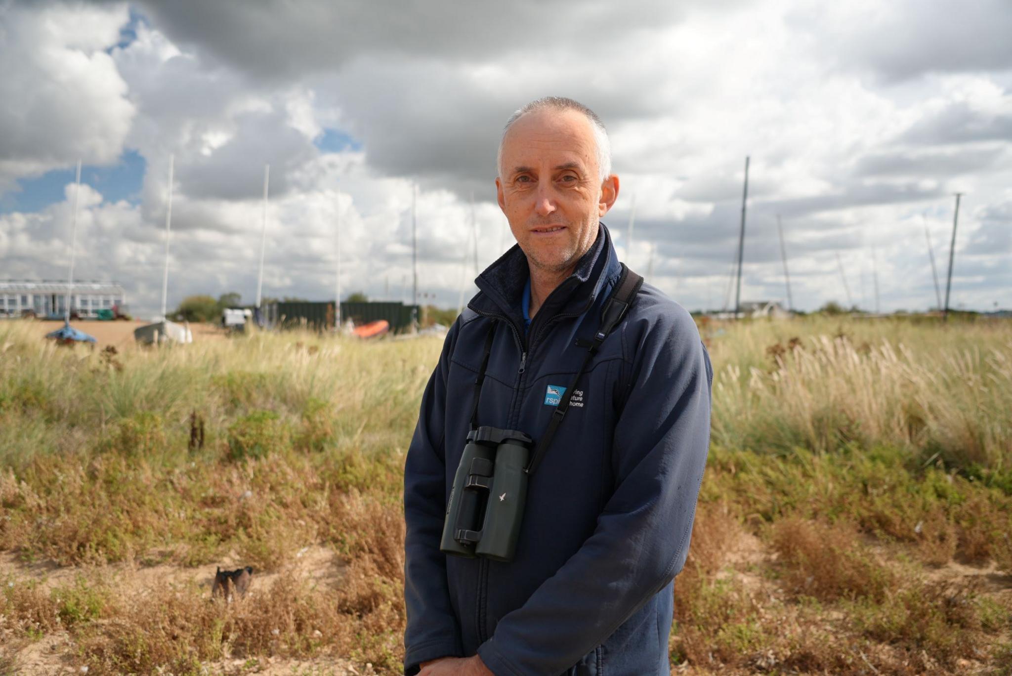 Man wearing dark-coloured RSPB jacket with binoculars round his neck standing amid tall and scrubby grass with boats in background