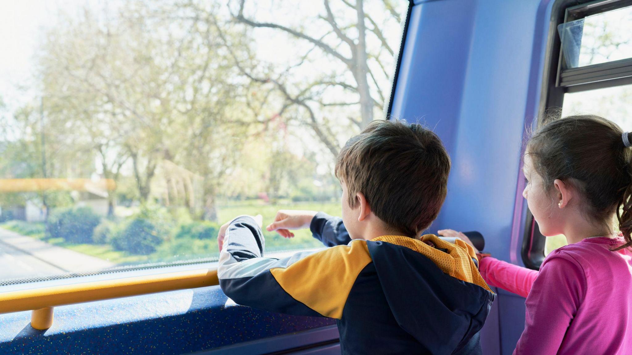 A boy and girl look out the front of a bus at a green field with trees.