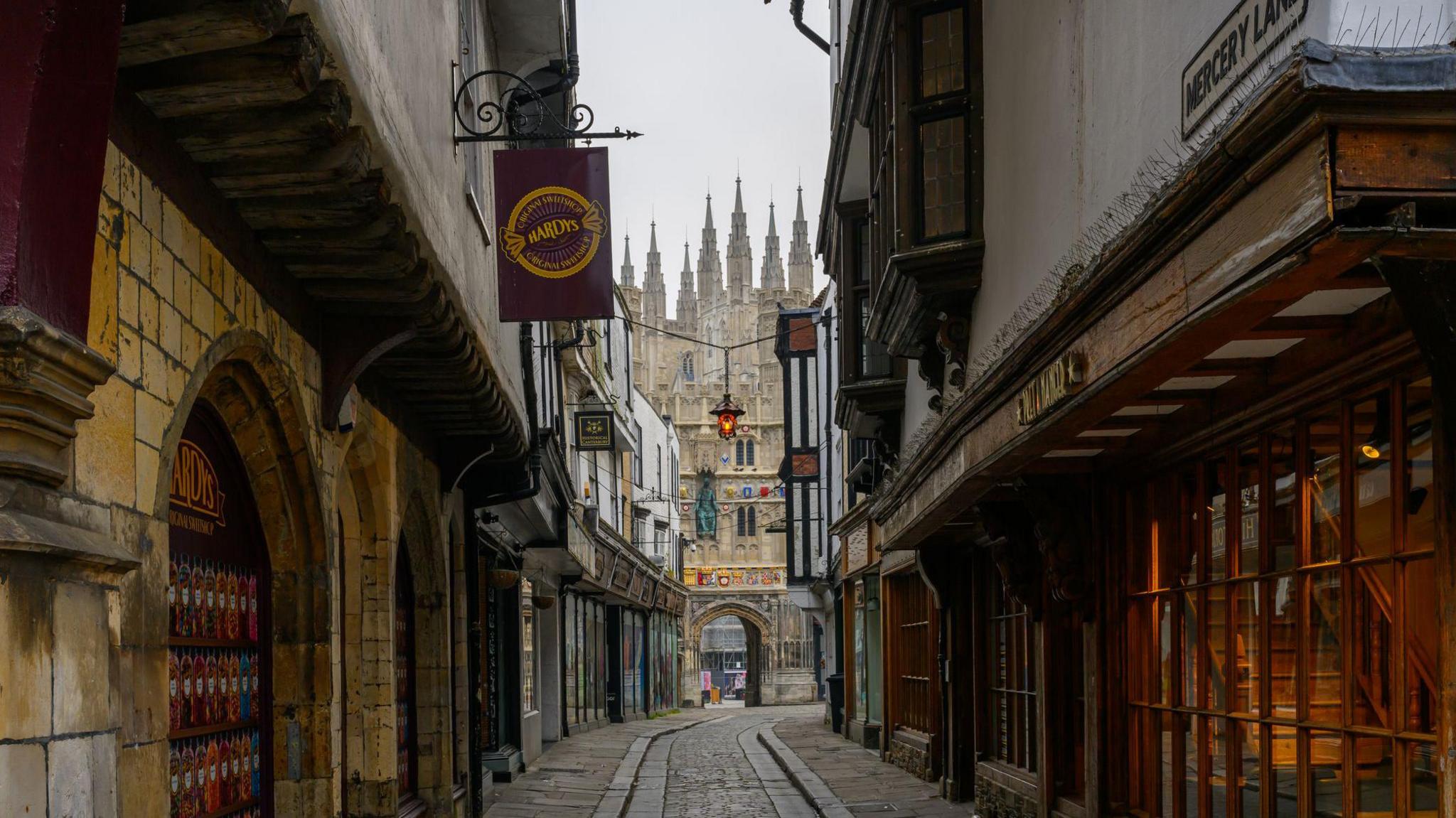 A narrow cobbled road leading up to an English Gothic style cathedral. Modern shops with traditional fronts line the cobbled street.