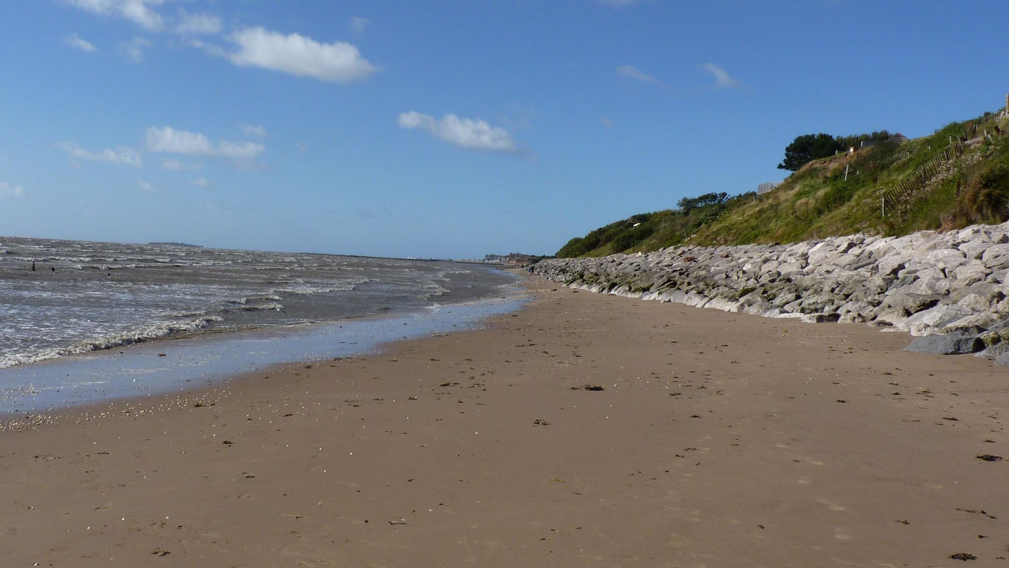 Waves splash on a sandy Wirral beach alongside rocks and a grassy slope under a blue sky, specked with clouds.